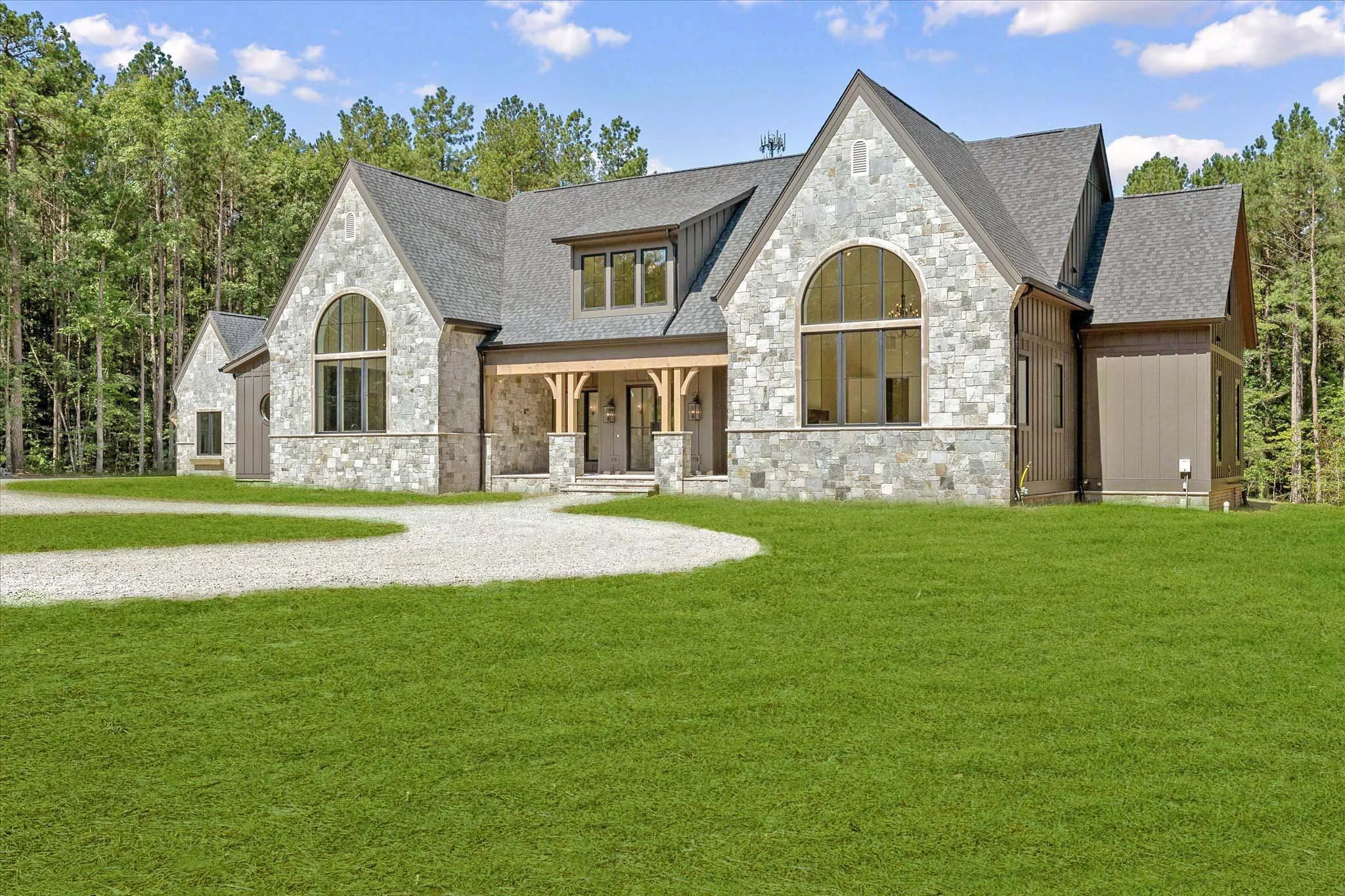 A large, modern house with stone and wood exterior surrounded by a grassy lawn and wooded area in the background.