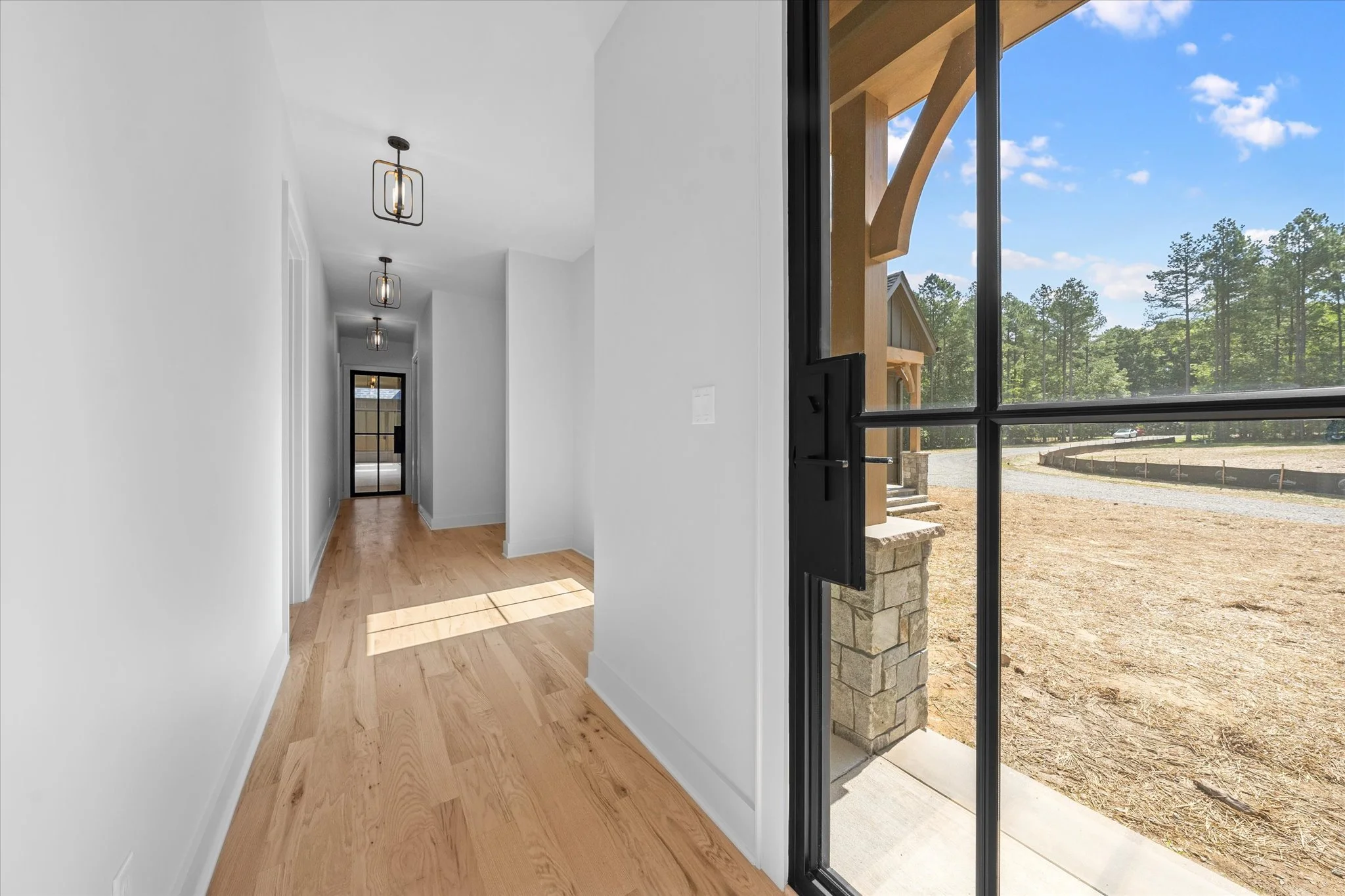 View of a hallway inside a house with light wood flooring, white walls, and modern ceiling lights, with a glass door at the end opening to an outdoor area with a dirt yard and trees.