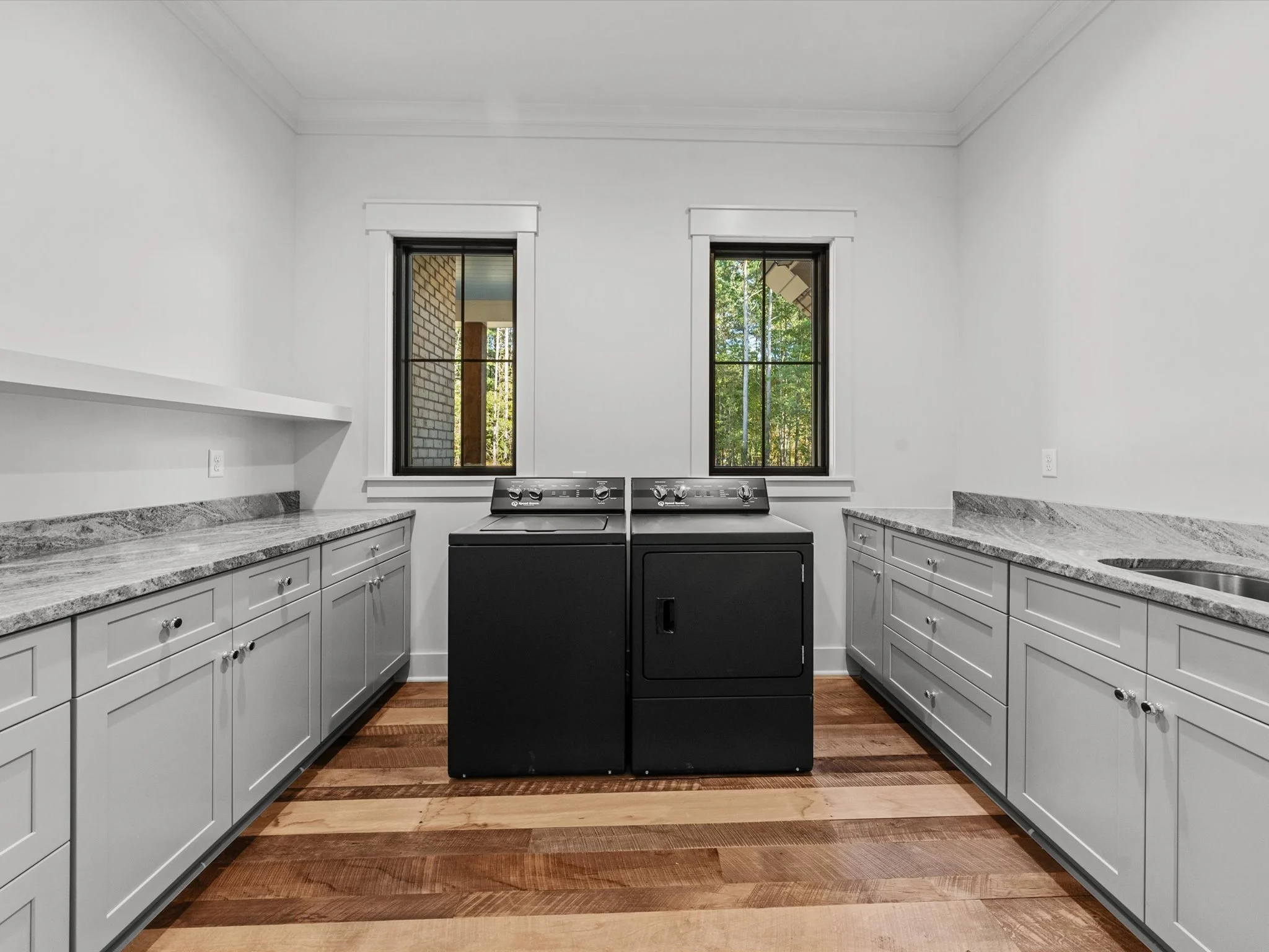 Laundry room with white cabinets, granite countertops, two black top-loading washing machines, and two windows showing greenery outside.