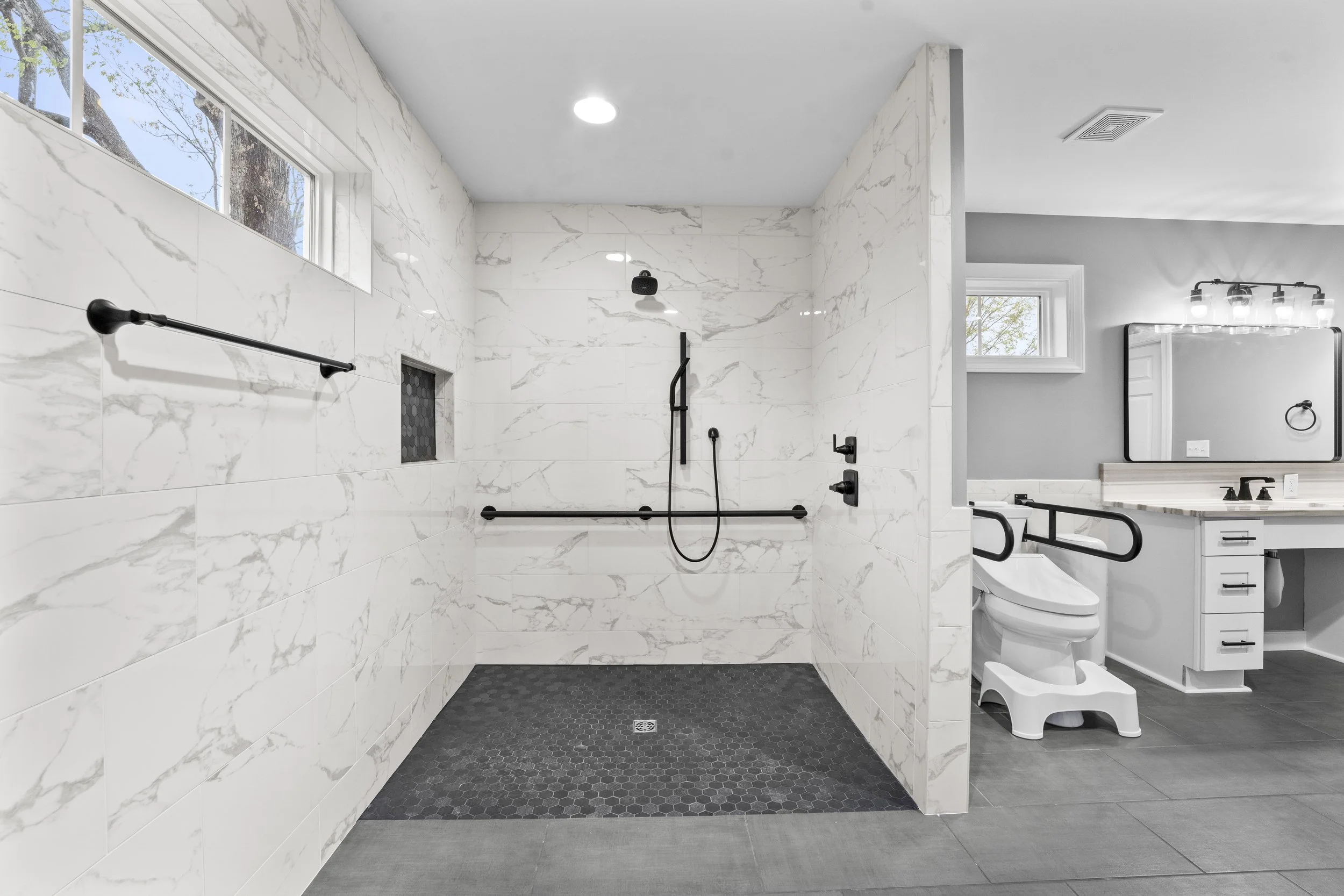 A spacious walk-in shower with white marble walls, black fixtures including a showerhead and grab bars, and dark hexagonal tile flooring.