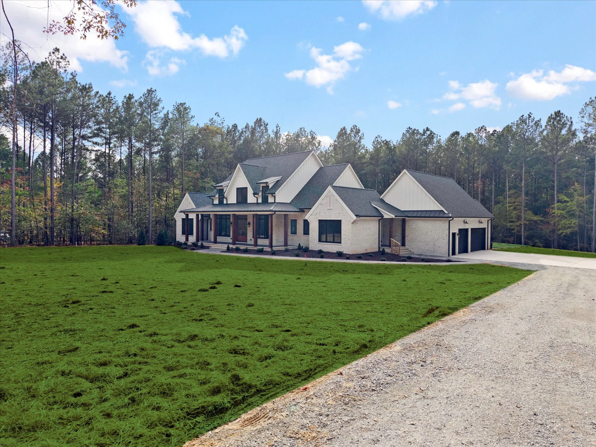 A large modern house with multiple gabled roofs, white brick exterior, and a porch, situated on a spacious grassy lawn with a gravel driveway and surrounded by trees under a partly cloudy sky.
