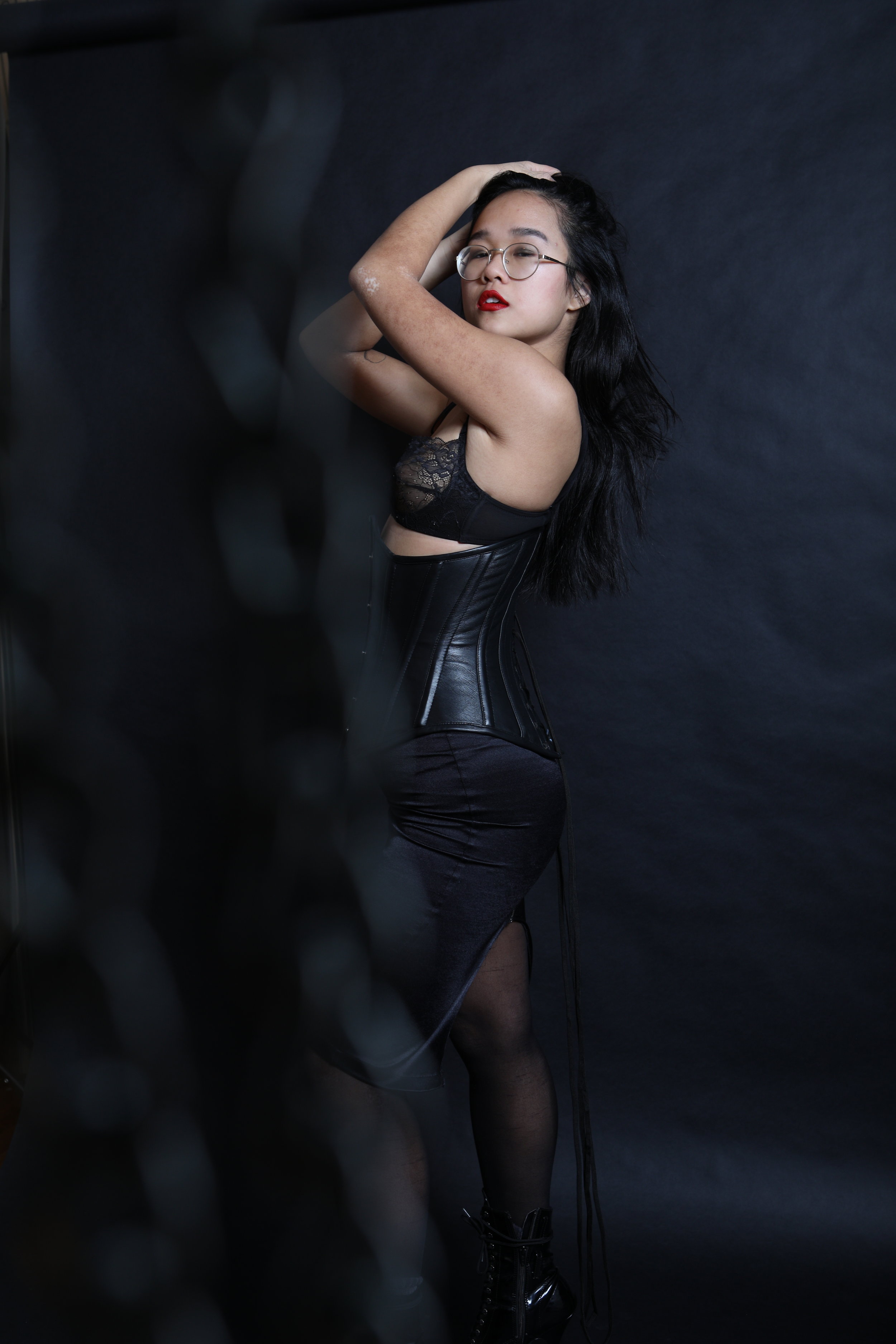 Chinese New York dominatrix and fetish lover Empress Wu lifts up her hair to reveal her outfit of lace lingerie, black leather corset, nylon stockings, and black vinyl boots.