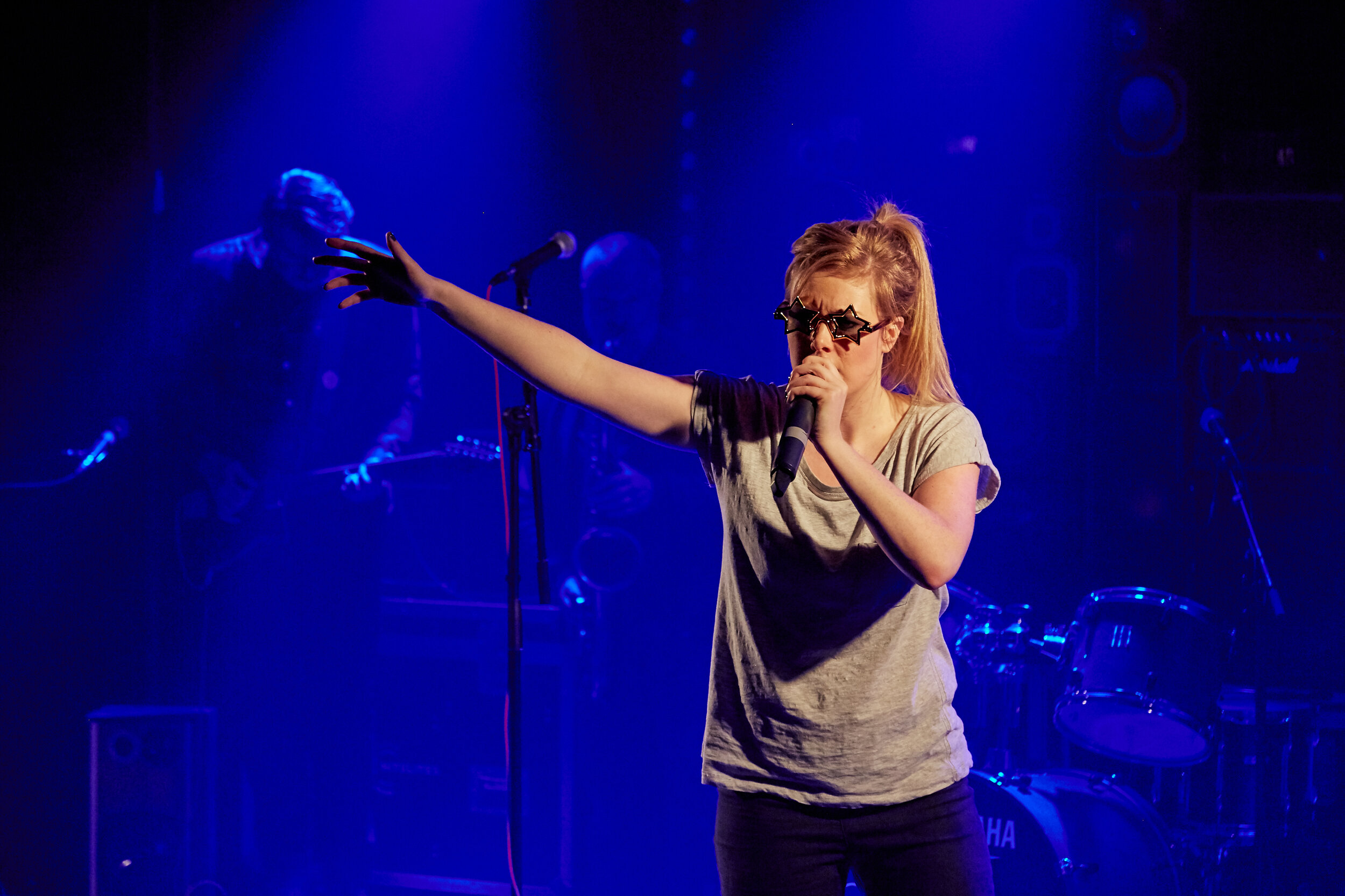  A singer wearing sunglasses holds a microphone to her mouth with an arm spread out in expression. 