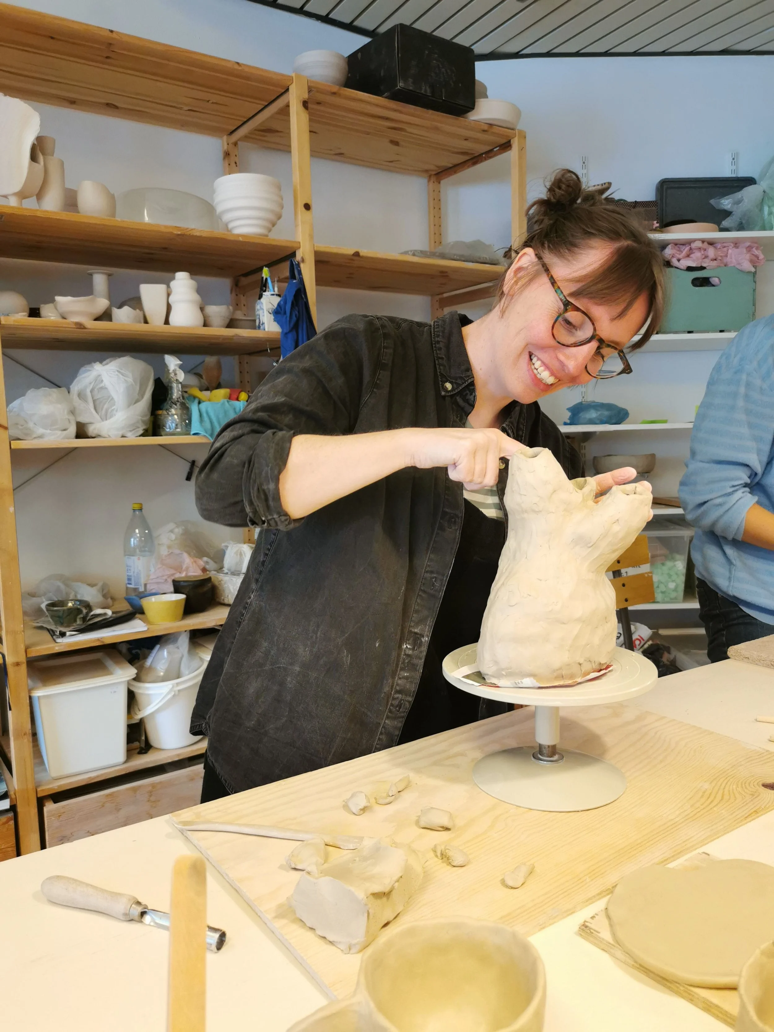 Rosie Priest, wearing a black shirt, glasses, and hair in a bun sculpting a clay object on a potters wheel in a well lit studio space.