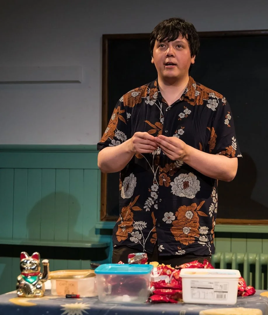 A man in a flowery shirt stands on stage behind a table containing various tupperwares, gazing outwards.
