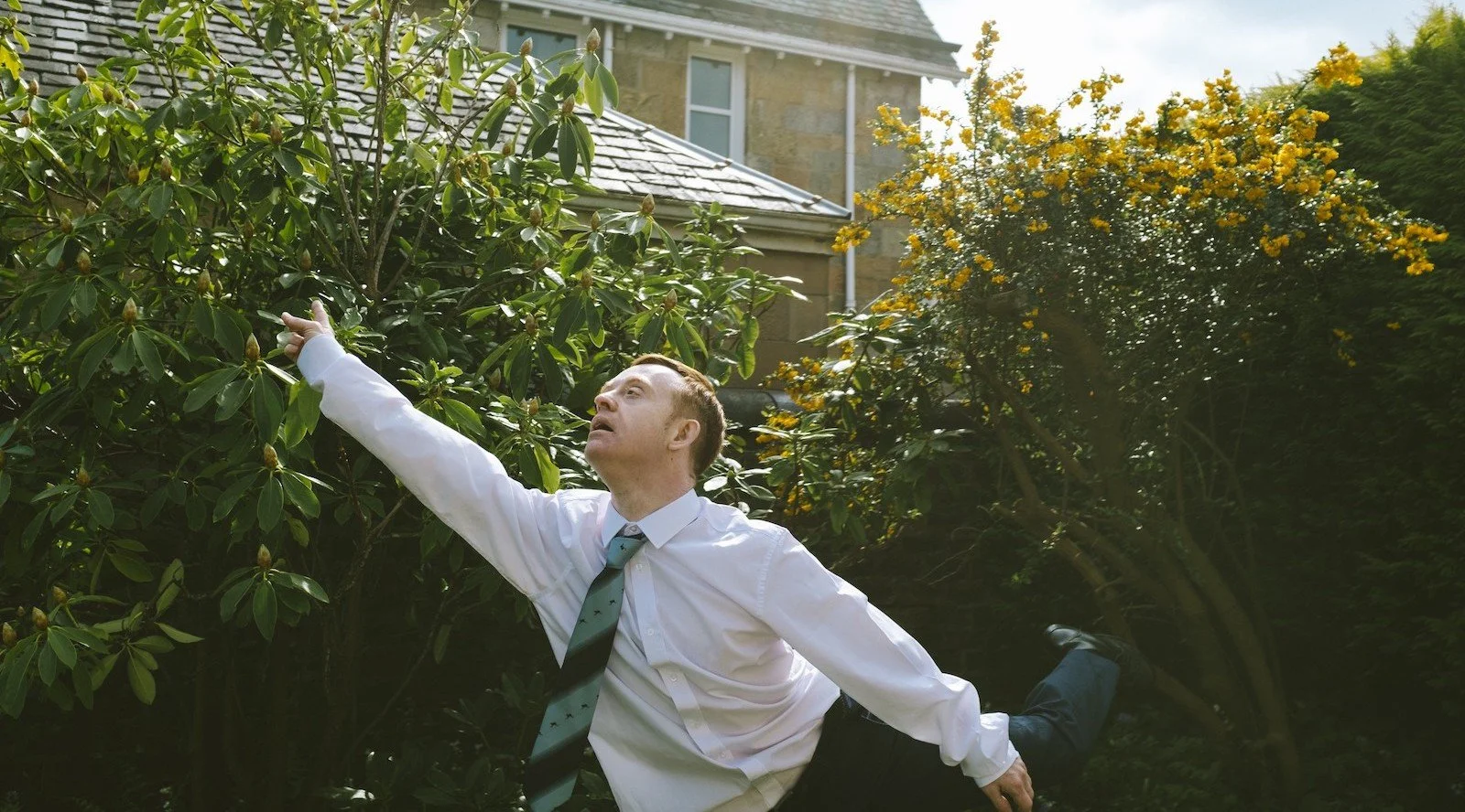 Neil, who has fair white skin, short ginger hair and is wearing a white shirt and blue striped tie, is pointing to the sky whilst lifting his leg behind him. In the background are dark green bushes, one with yellow flowers and part of a house.