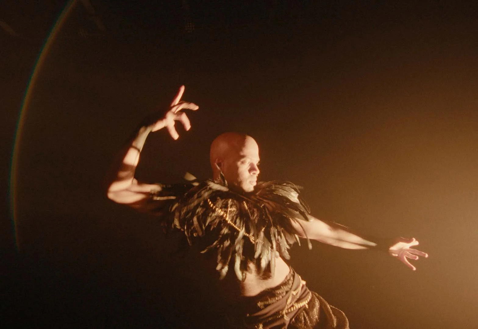 The performer is looking towards the light with tension in his hands. One hand is lifted and the other is stretched in front of him. He wears a black feather piece on his shoulders and black shorts.