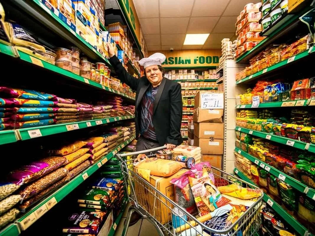A man in a chef's hat and black blazer stands in an aisle of food shop with a full trolley, reaching towards the top shelf.