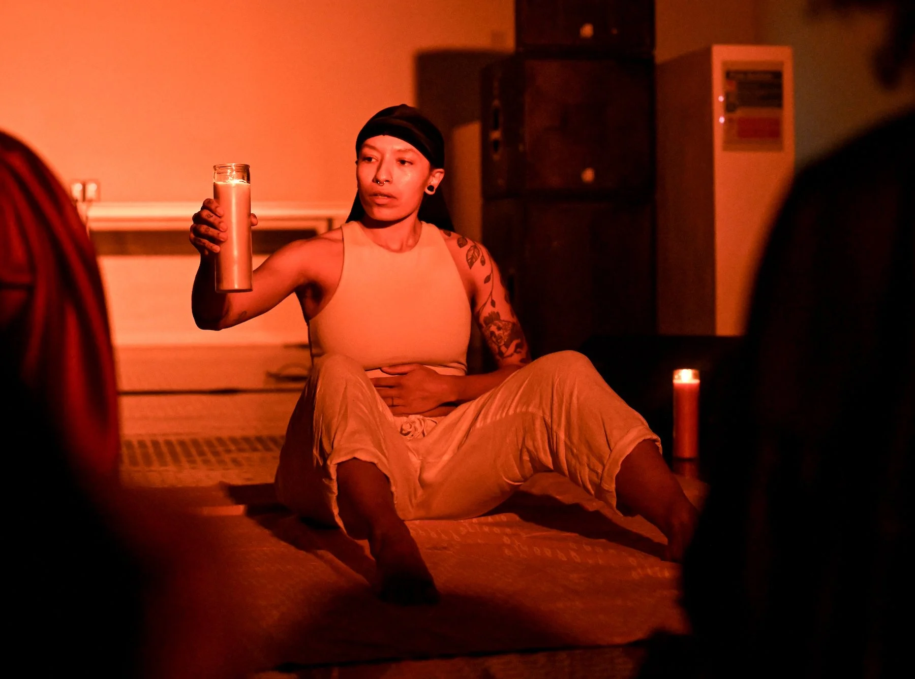 A brown trans masculine person is sitting on the ground wearing cream linen trousers, a cream binder and black durag. They are holding up a candle encased in glass and sitting under a warm red glow in front of an audience.