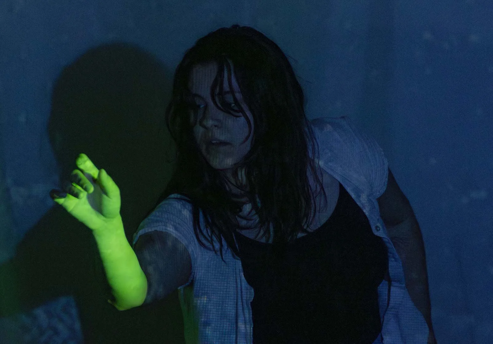 A performer with one arm extended in front of her, her arm lit in a bright green light, the rest of her body lit in deep blues with textures of water rippling.
