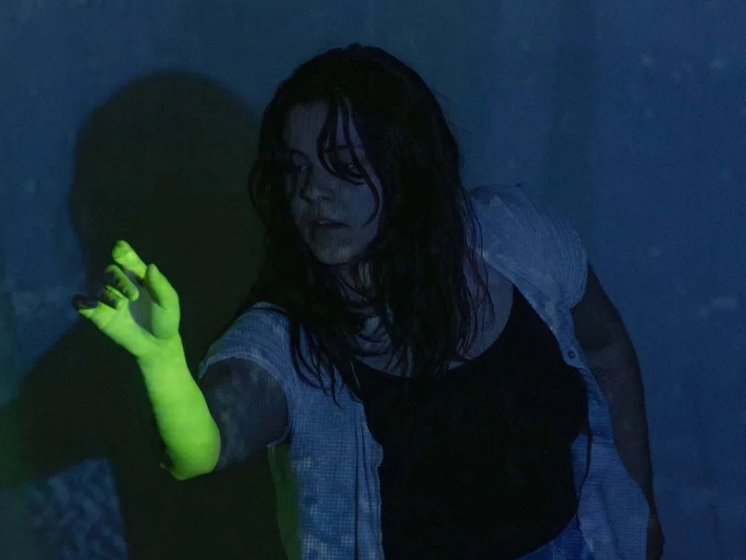 A performer with one arm extended in front of her, her arm lit in a bright green light, the rest of her body lit in deep blues with textures of water rippling.