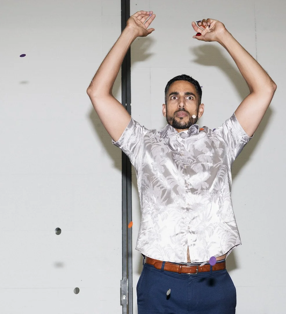 A performer stands with arms raised dramatically above his head, eyes wide, in a white room. Small pieces of coloured confetti are suspended mid-air around him.