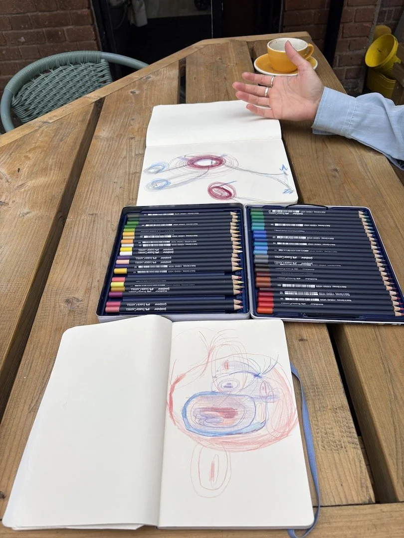 Two Sketch books with abstract drawings laid out on a wooden table. There is also a tin of colored pencils open in between the two sketchbooks. At the top right is a human hand can be seen in an open gesture.