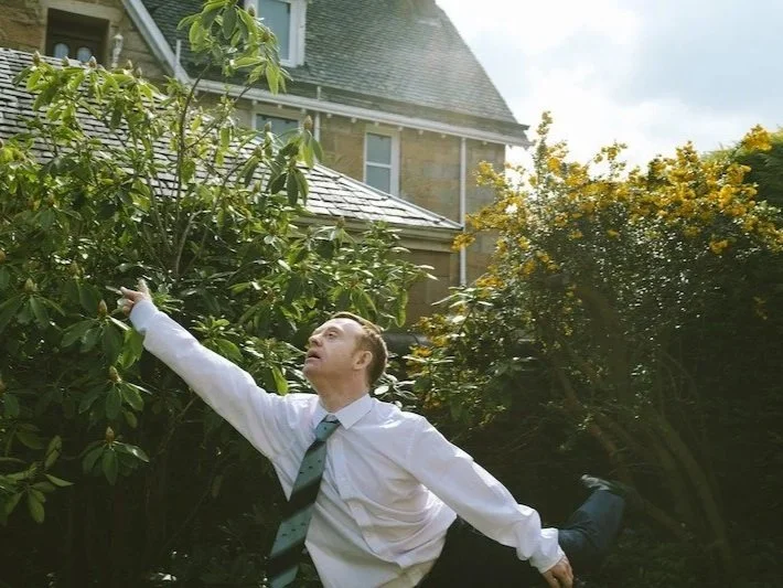 Neil, who has fair white skin, short ginger hair and is wearing a white shirt and blue striped tie, is pointing to the sky whilst lifting his leg behind him. In the background are dark green bushes, one with yellow flowers and part of a house.
