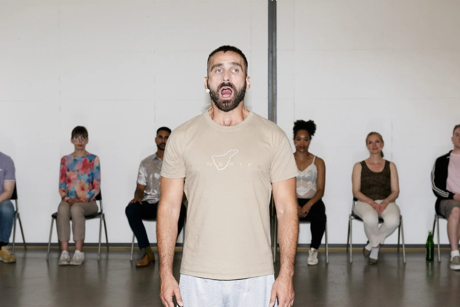 Image 3 A performer stands at the front, singing with mouth open and eyes focused. Behind him, six seated performers look on from folding chairs. The group wears casual clothing and headset microphones in a bright room.