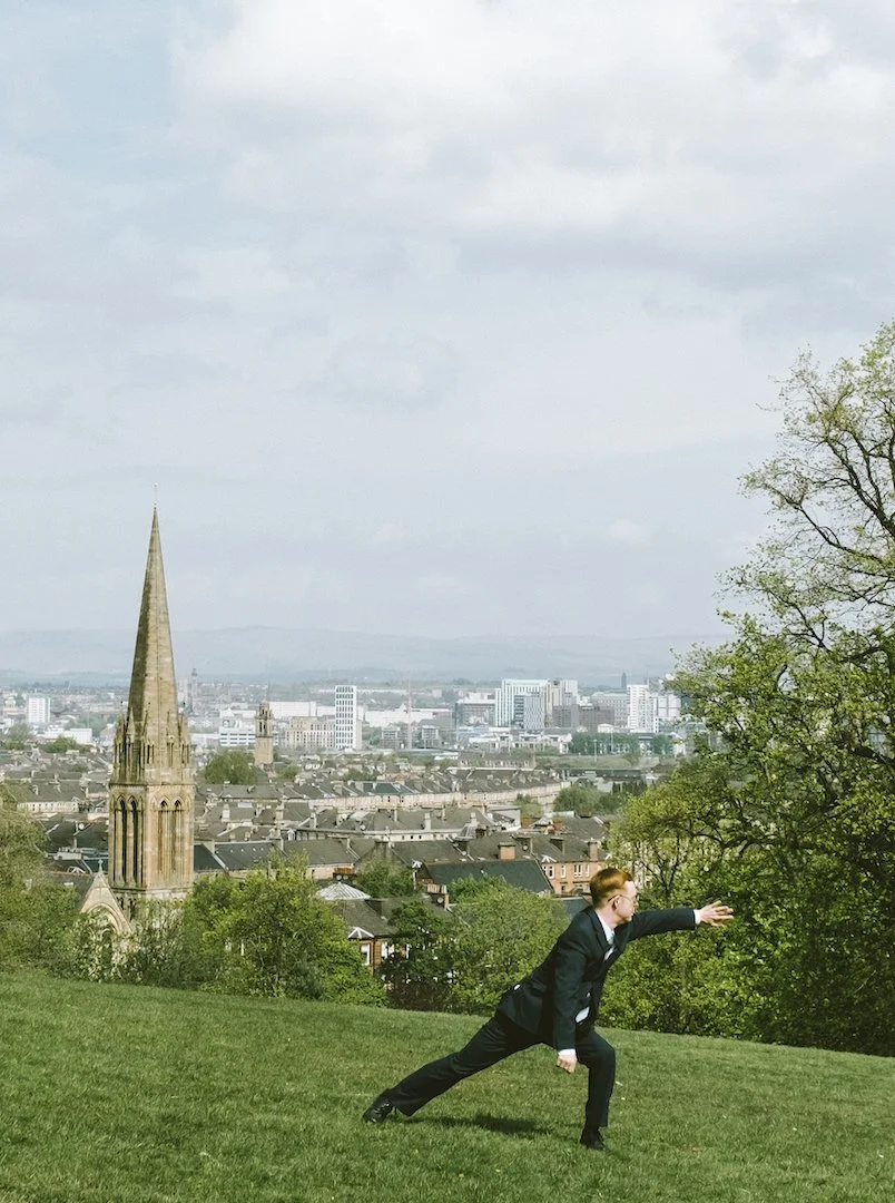 Neil, who has fair white skin, short ginger hair and is wearing a dark blue suit, is reaching to the side, whilst standing on a grassy green slop in Queens park, with a view of the city behind him.