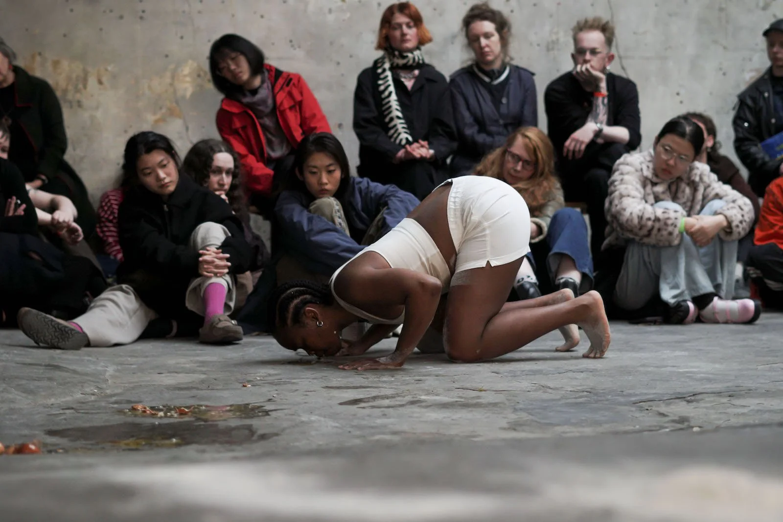 n:u (melissandre varin), a dark skinned person with braided hair on their hands and knees kissing the ground which has eggs and flour on it. They wear white shorts and a vest. Behind them is a small crowd of audience members watching intently.