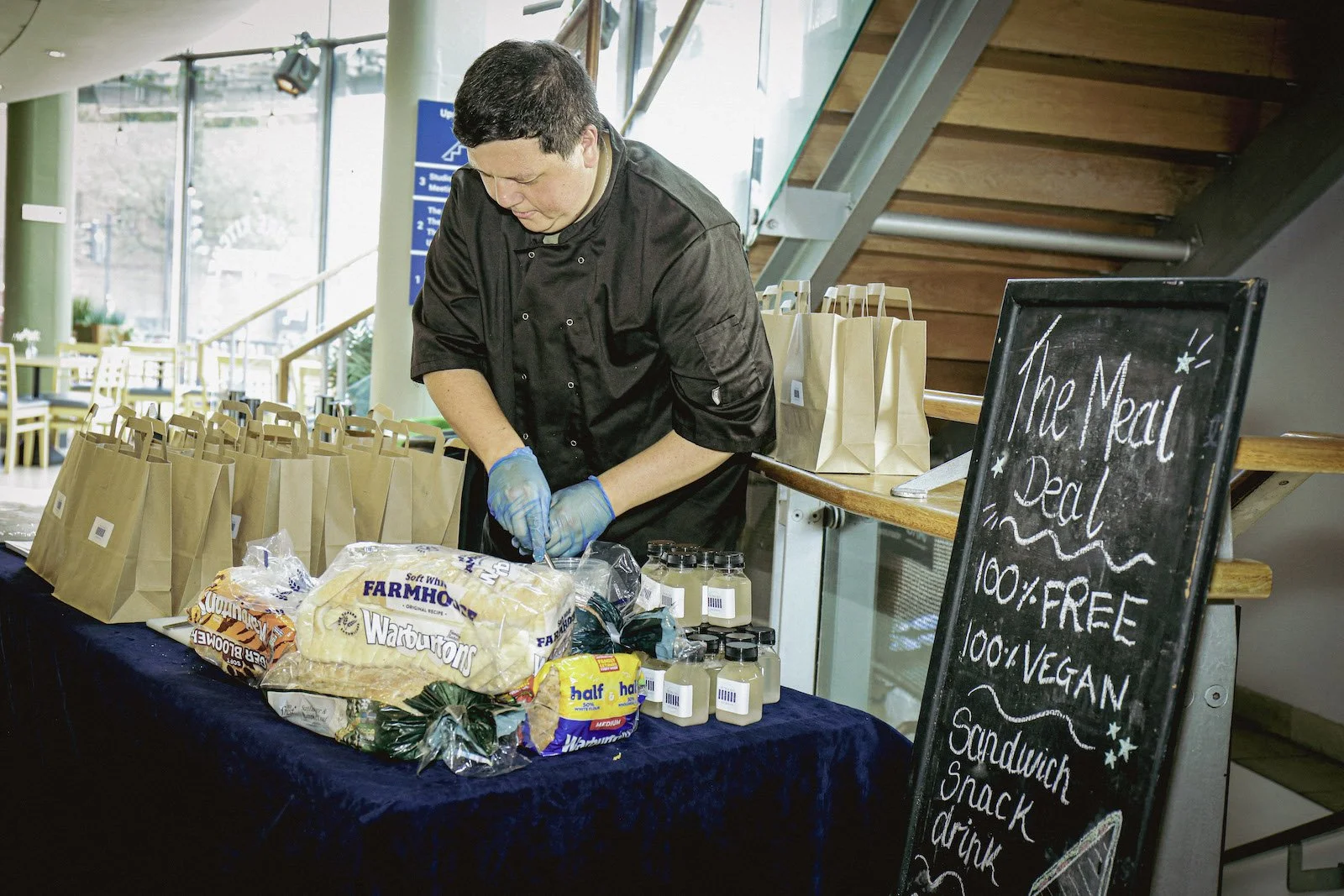A man in a chef uniform is behind a table with various food items, wearing blue kitchen gloves and cutting something.
