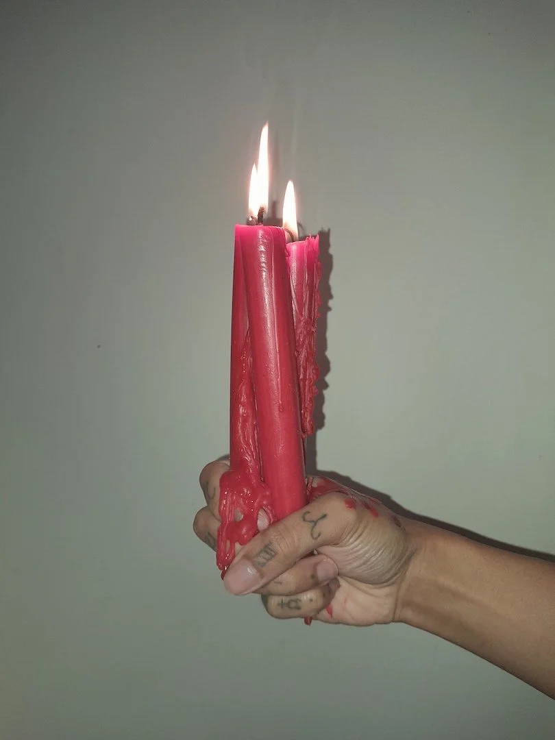 A brown hand with astrological finger tattoos holds three red taper candles. The candles are melting wax over the hand's fingers.