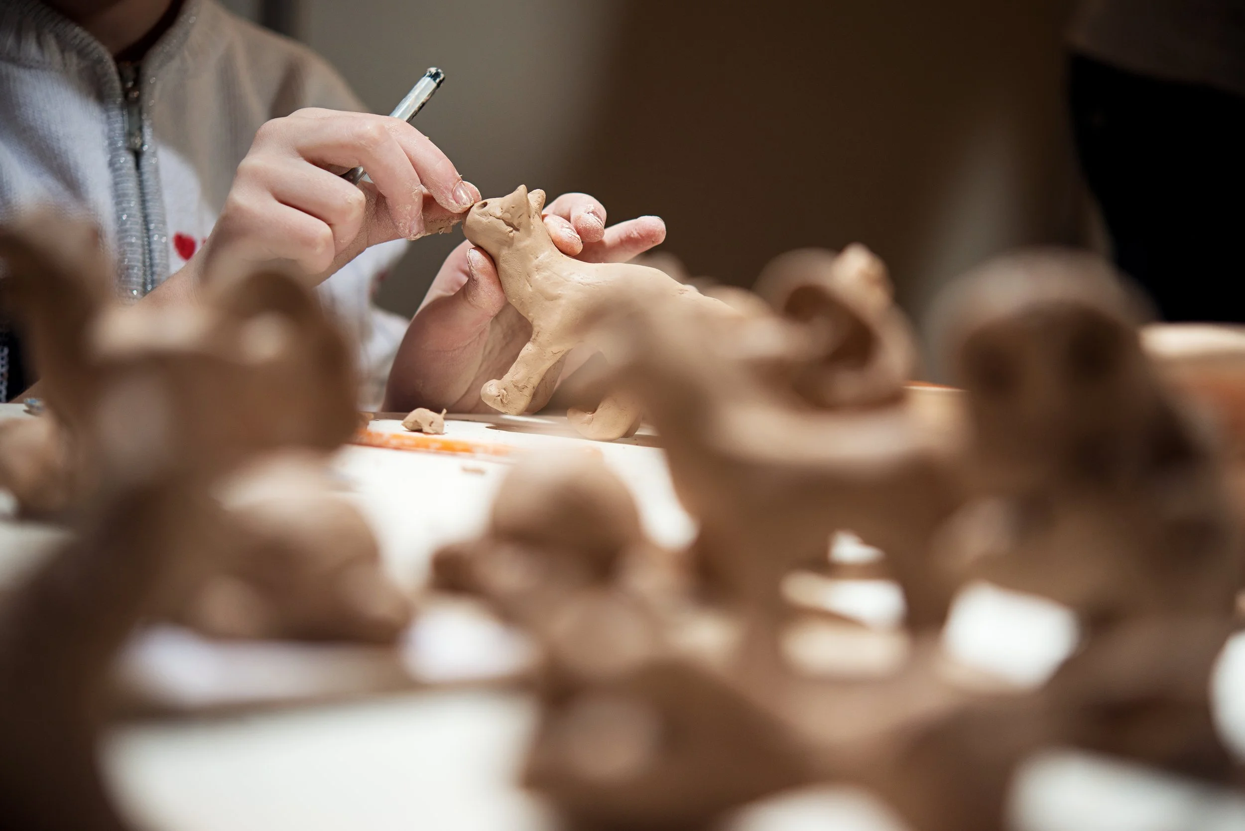 An image with a series of clay figurines of animals in the foreground, behind them you see Rosie Priest's hands making a small cat like model. The image feels warm with a lot of brown tones.