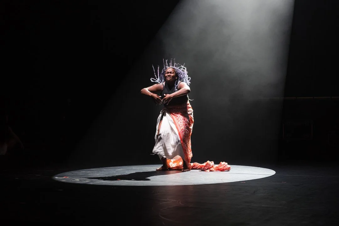 “A dancer with purple braids stands alone in a spotlight on a dark stage, wearing a patterned cloth wrapped around their waist. Her face is expressive her hair up in the air
