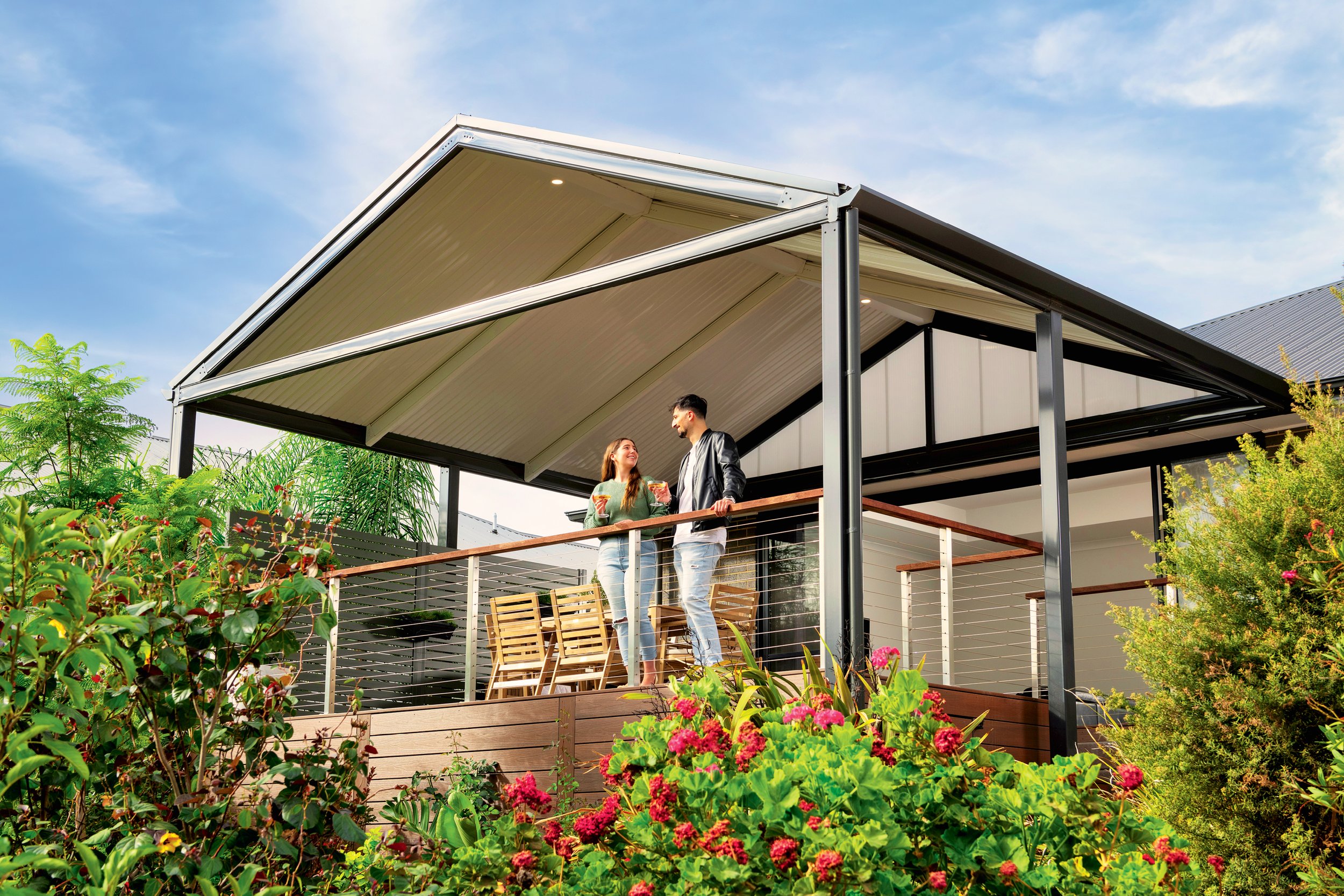 A modern house with a balcony and a couple standing and talking on it, surrounded by lush green plants and flowers, under a blue sky with some clouds.