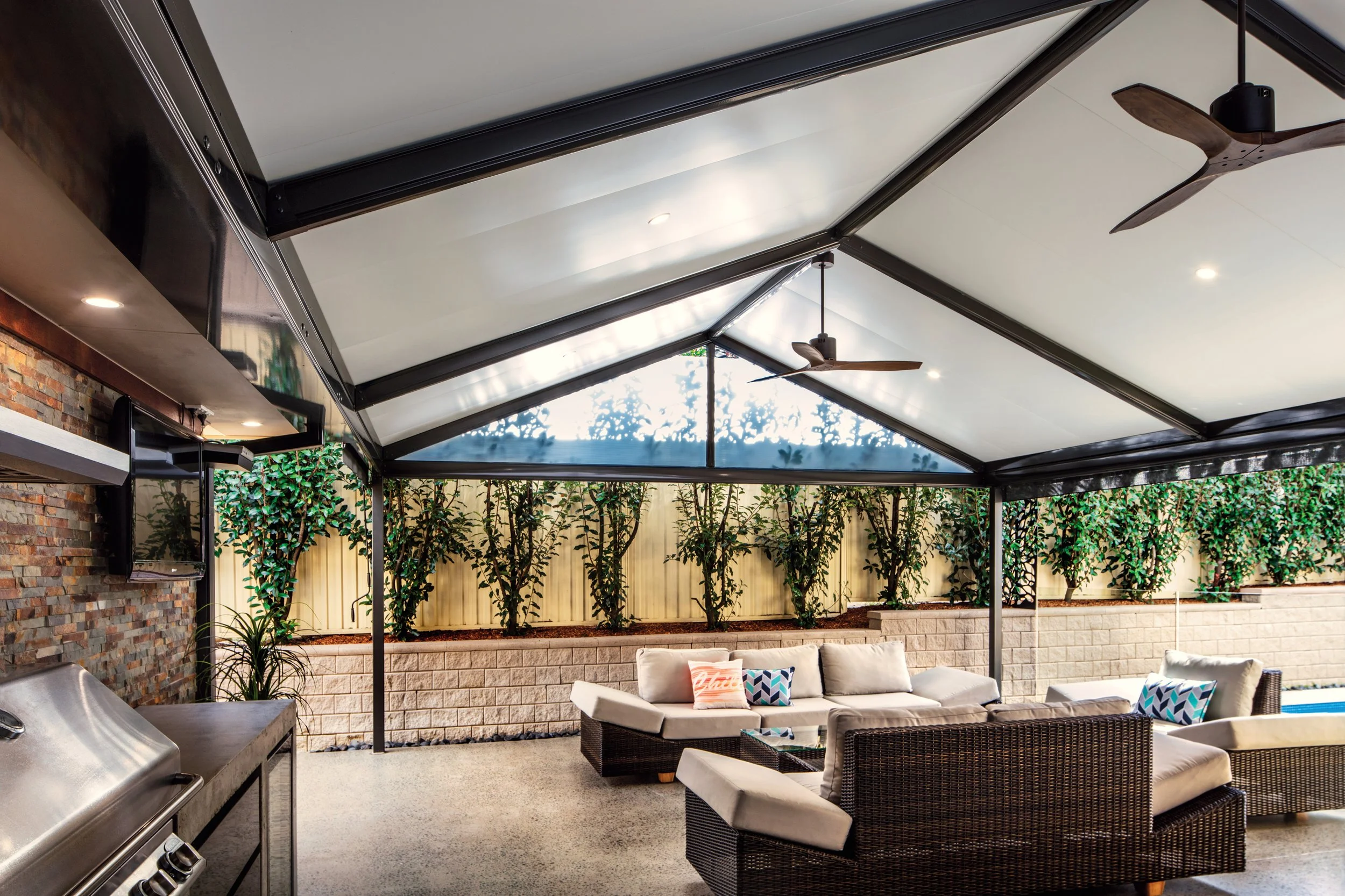 Covered outdoor patio with ceiling fans, string lights, wicker sofa with colorful pillows, built-in grill, and a wooden ceiling fan, enclosed by a beige fence and landscaped with small trees.