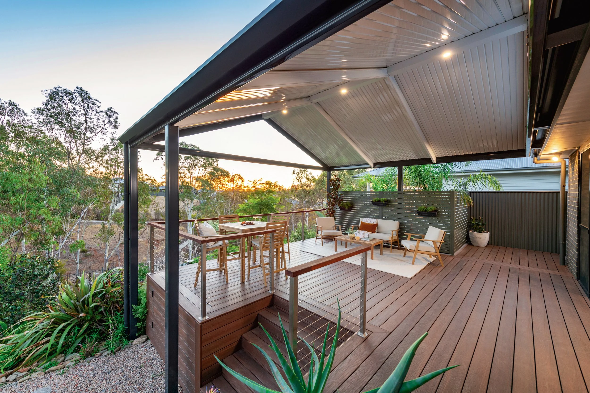 Outdoor patio with wooden deck, seating area, and a roof, overlooking trees and a sunset sky.