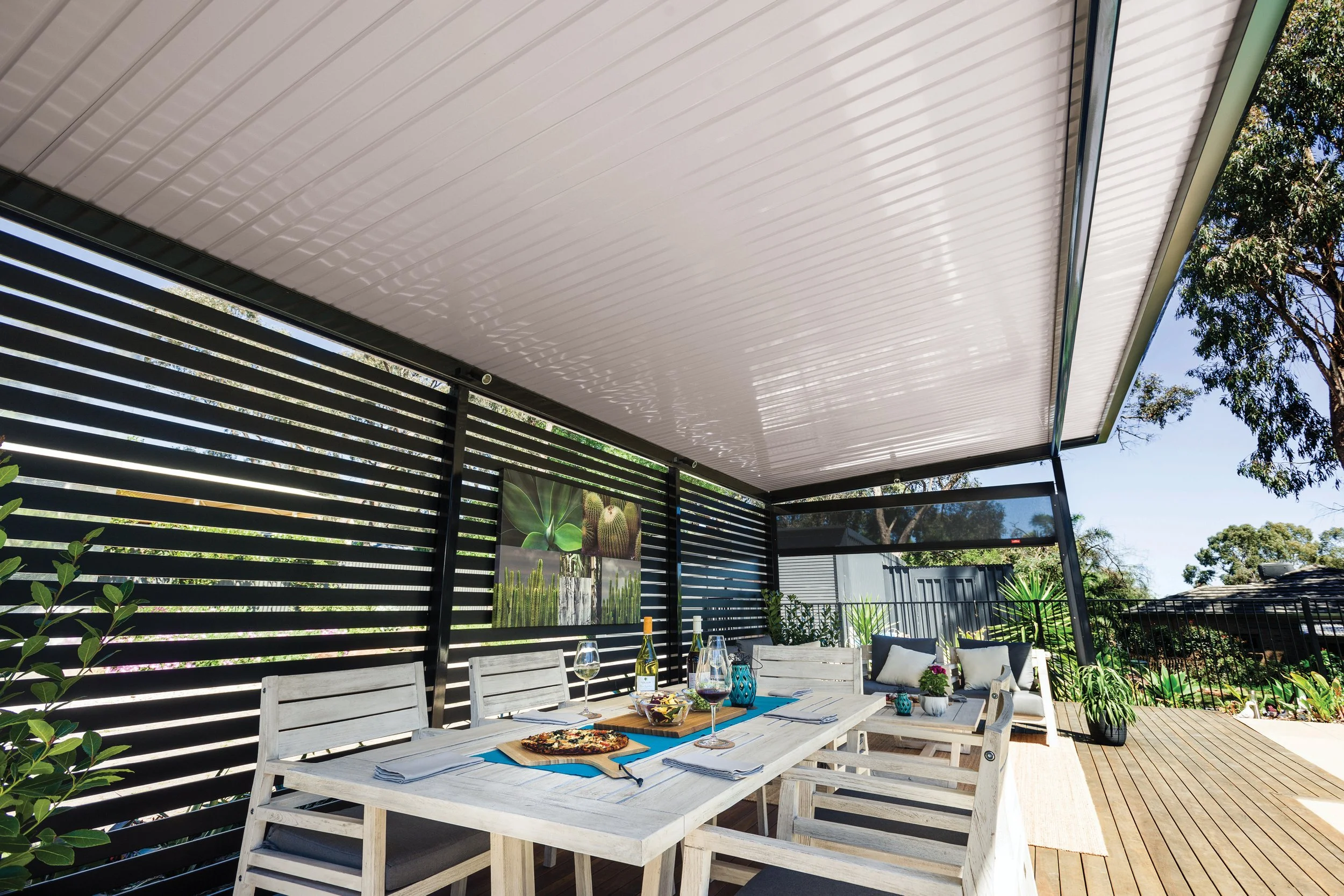 A modern outdoor patio with a white wooden table set for a meal, surrounded by matching chairs, under a white pergola with a shaded roof, with greenery and trees in the background.