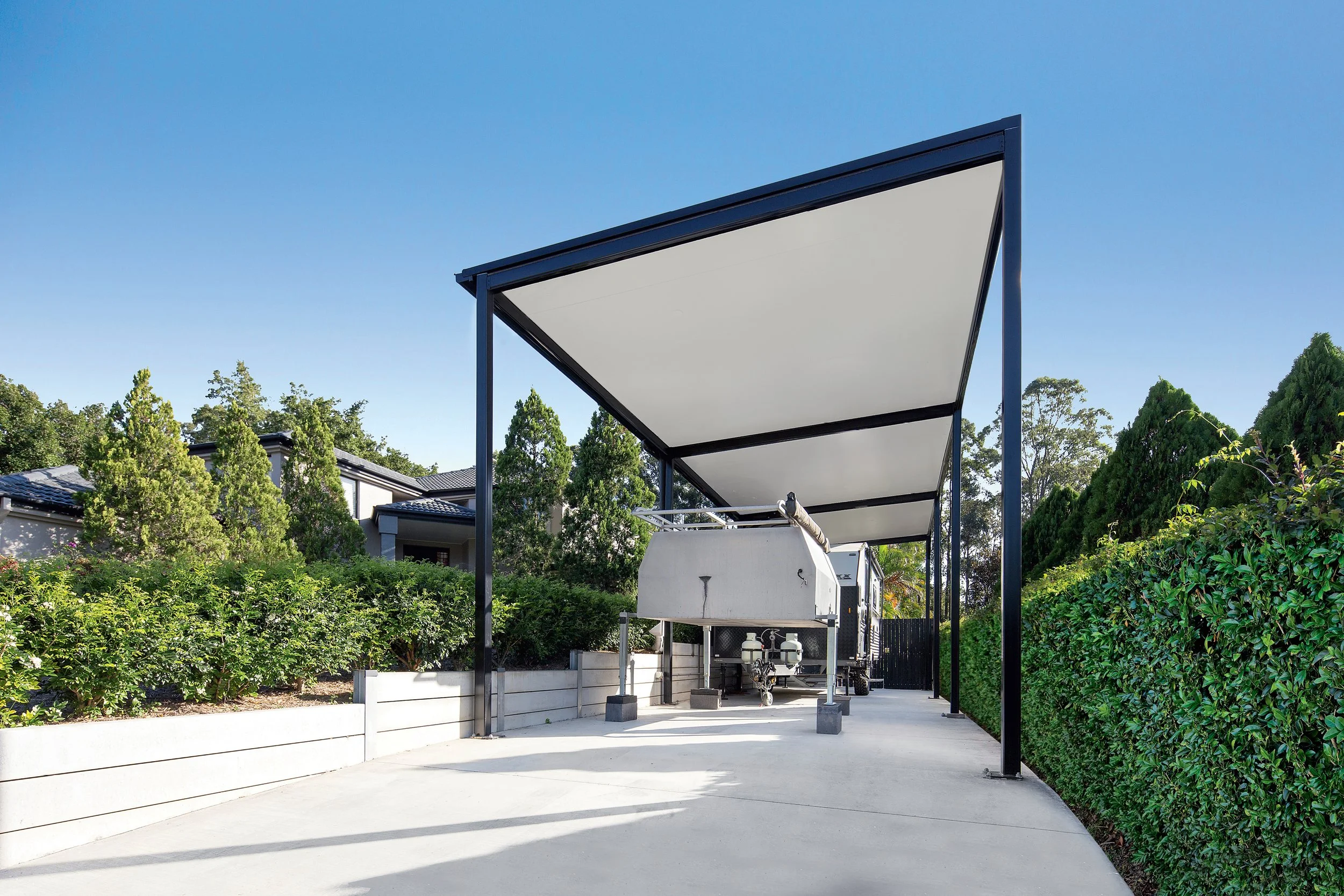 A modern shade structure with a white canopy supported by black metal poles, situated on a concrete patio surrounded by lush green shrubbery and trees, with residential houses in the background under a clear blue sky.