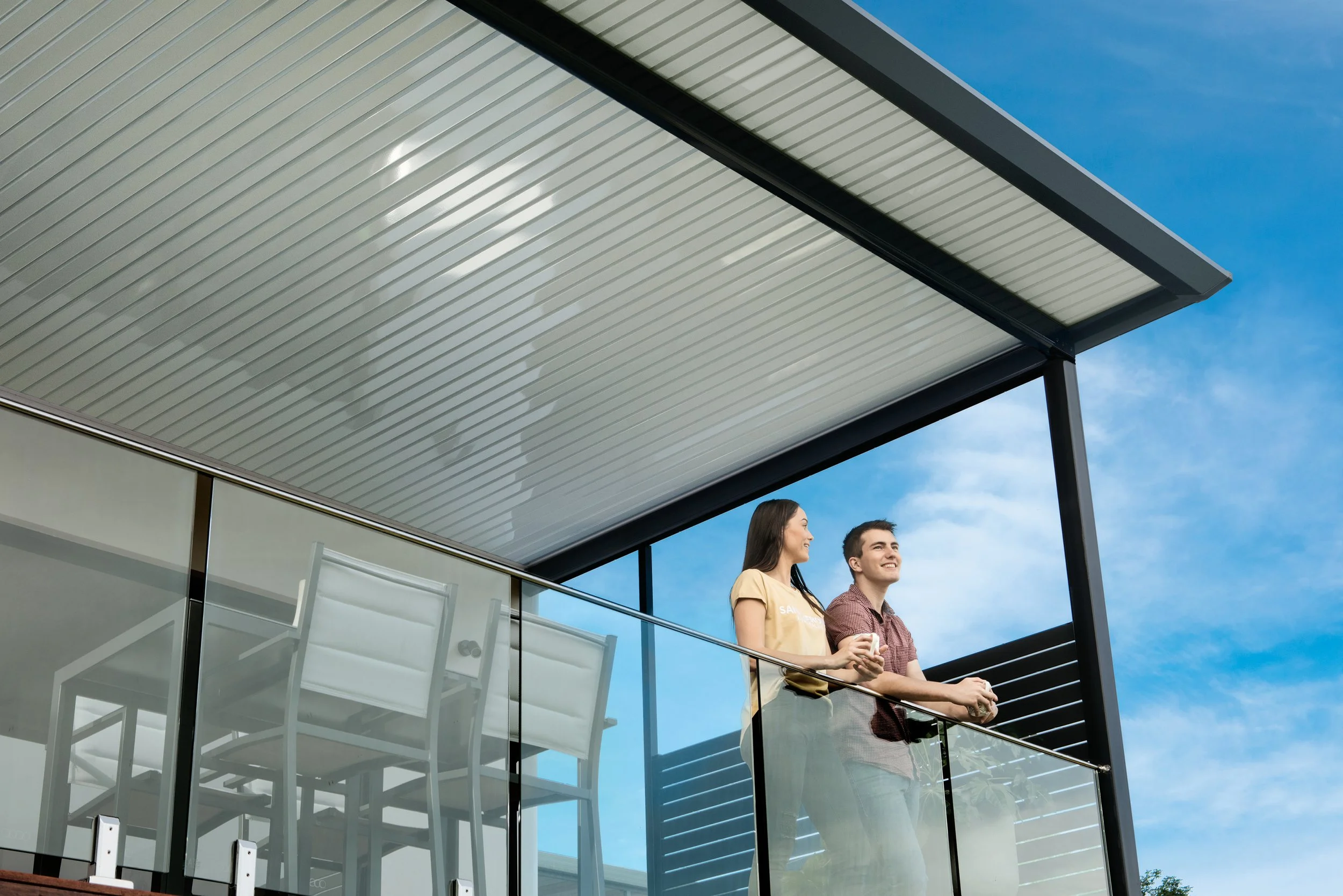 Two people, a woman and a man, standing on a balcony of a modern building with glass railing, smiling and looking out into the distance during daytime with a blue sky in the background.