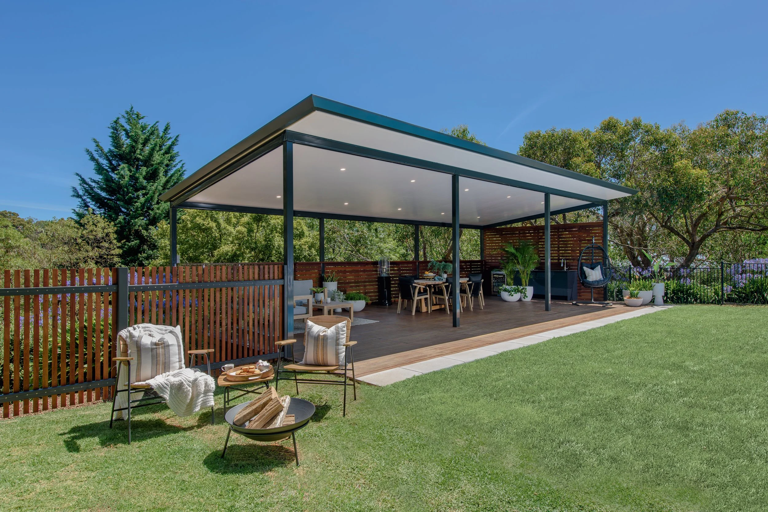 Modern outdoor patio with a covered seating area, surrounded by trees and a grassy lawn, with chairs, tables, and decorative plants.