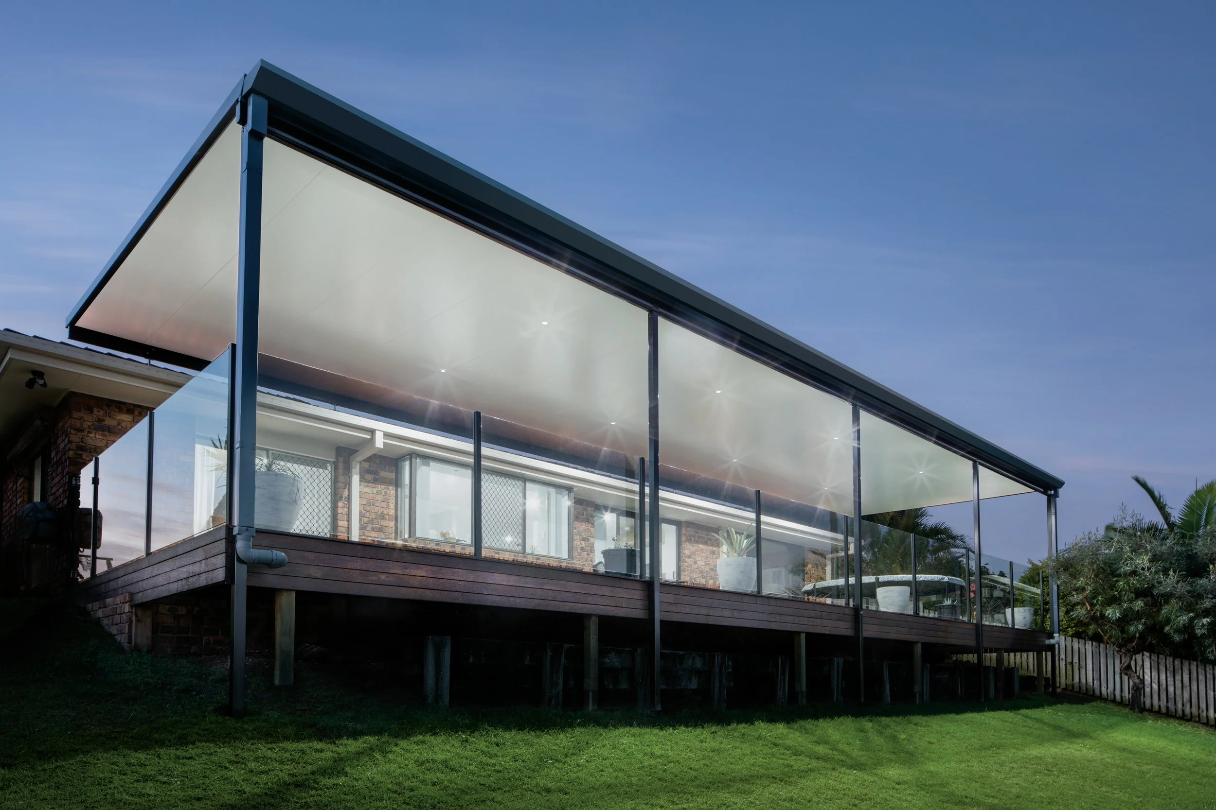 Modern house with a glass balcony and a white roof, set against a clear blue sky, with a grassy yard below.