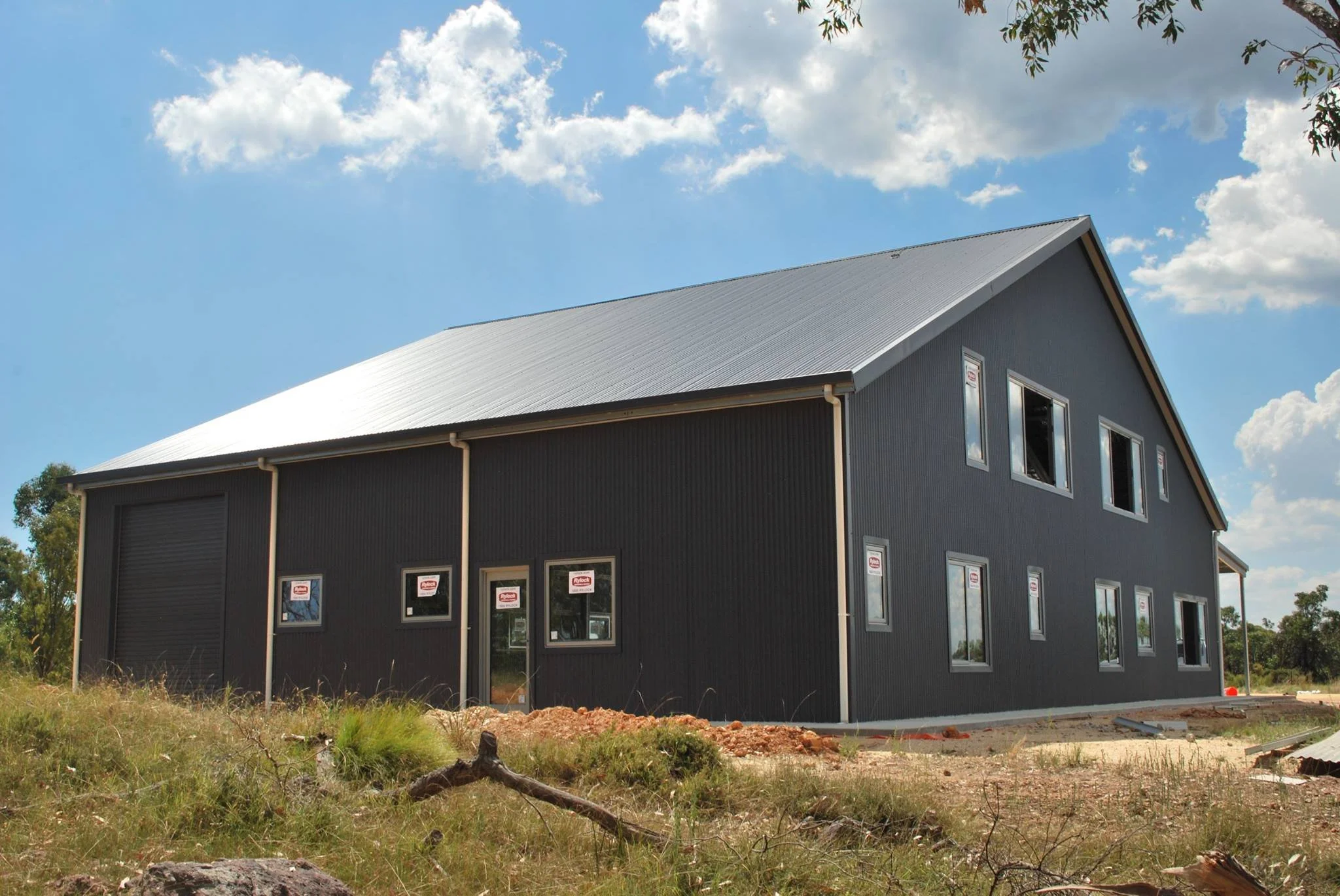 A two-story house under construction with dark exterior siding and multiple large windows, set against a partly cloudy blue sky with trees around.