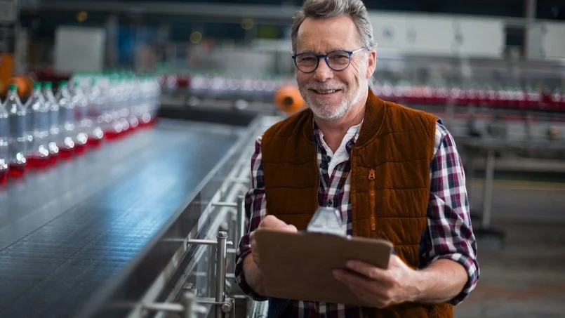 A man with glasses, a beard, wearing a plaid shirt and brown vest, smiling and holding a clipboard in an industrial or brewery setting.