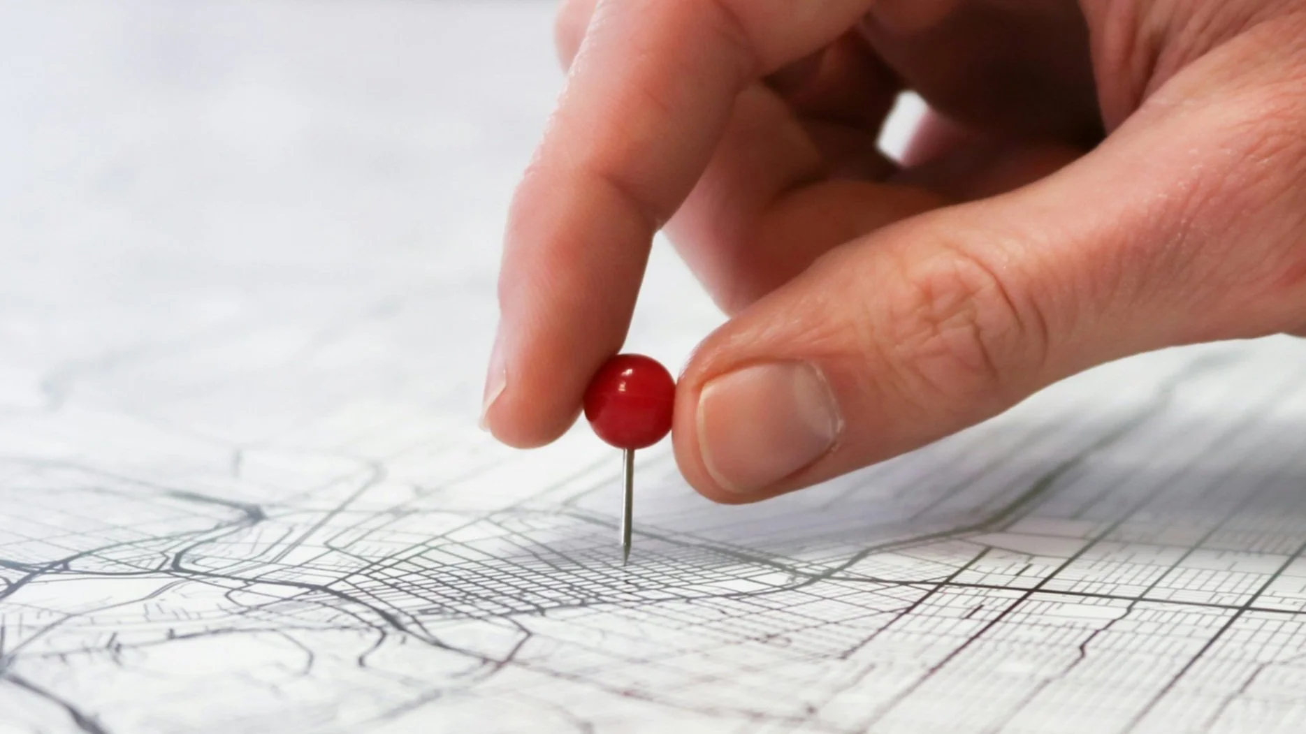 A hand holding a red pushpin, pinning a paper city map.