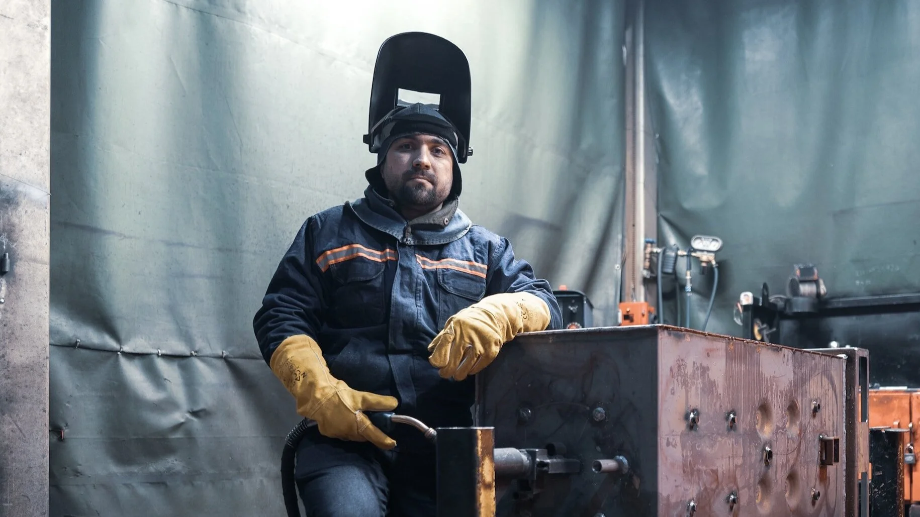 A welder with a welding helmet raised, dressed in protective gear and gloves, standing next to a large metal structure in an industrial workspace.