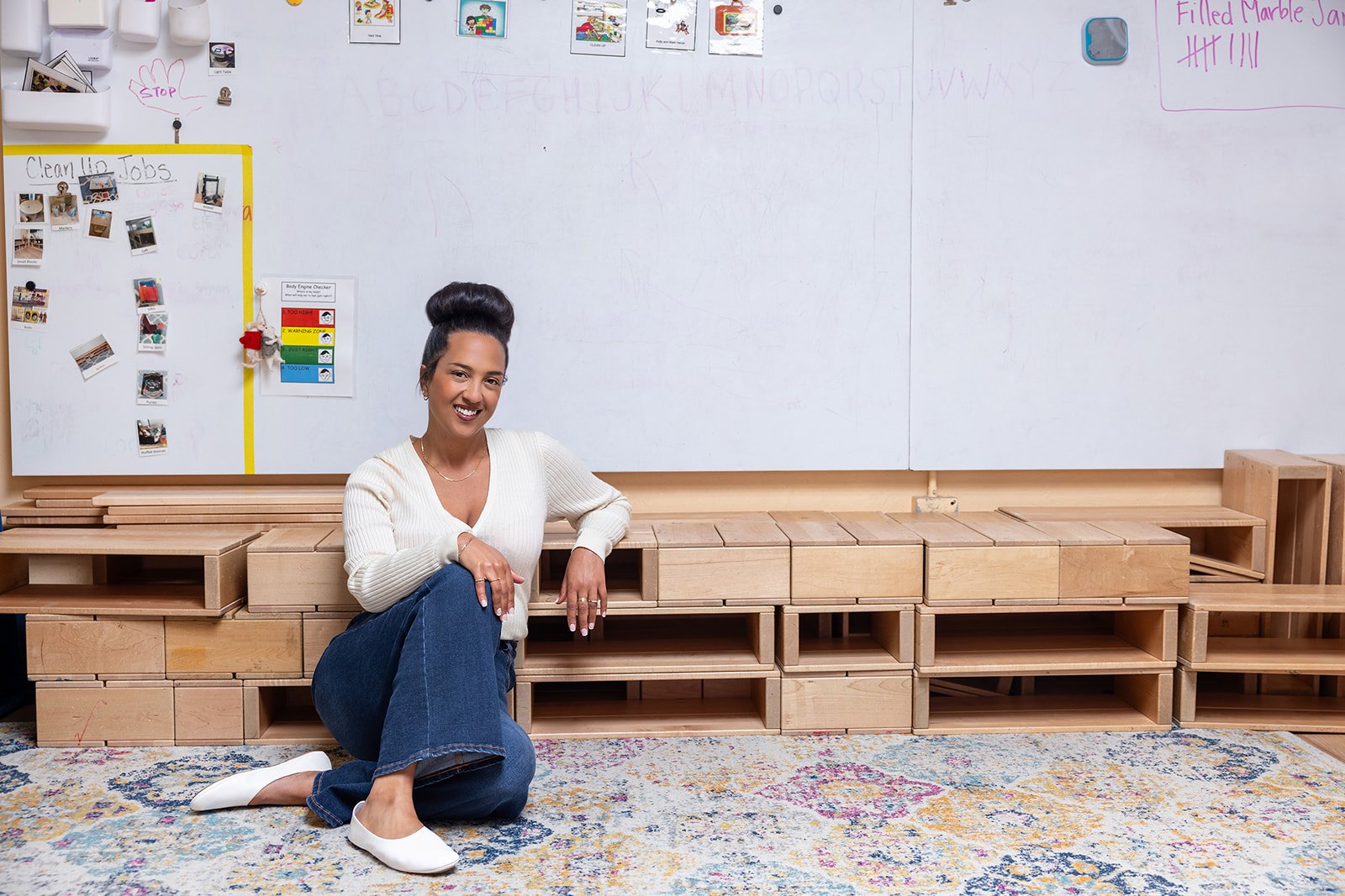 A woman posing and smiling in front of a shelving unit made of wooden blocks in a classroom or craft room, with a whiteboard and colorful decorations on the wall behind her.