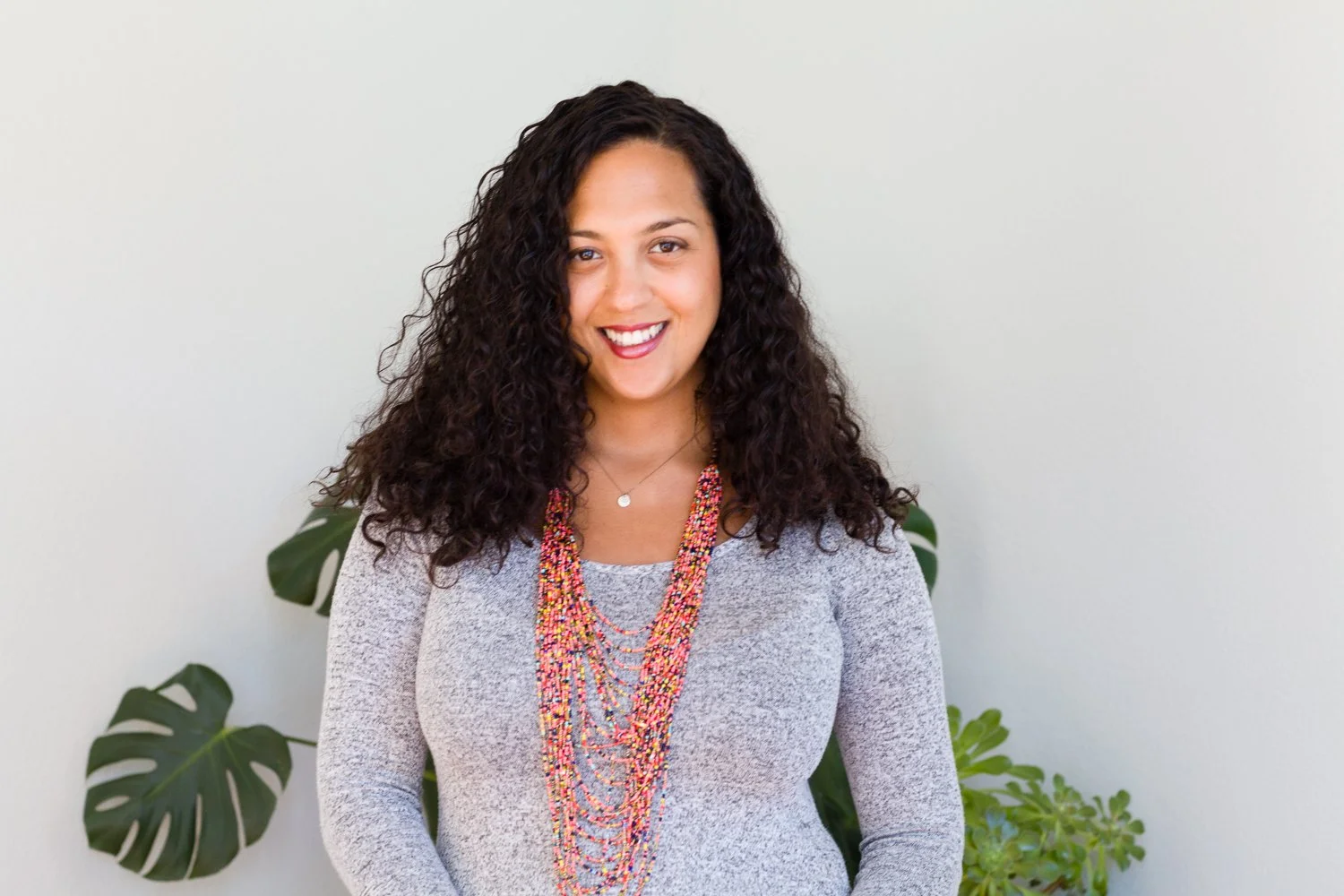 A smiling woman with curly dark hair wearing a grey sweater and colorful beaded necklaces standing in front of a plain white wall with green plants.