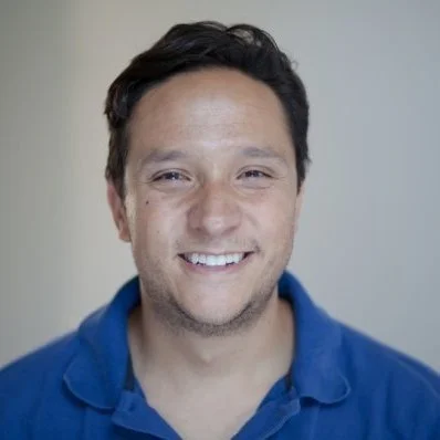 A smiling man with dark hair, wearing a blue collared shirt, standing against a plain light-colored background.