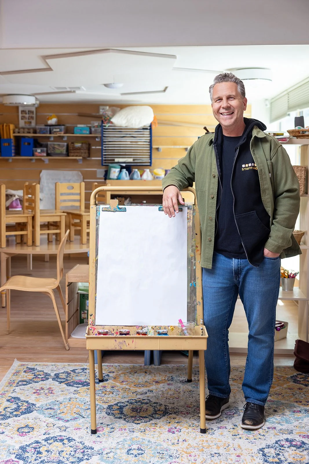 A smiling man casually dressed in a green jacket and blue jeans standing next to a small whiteboard on an art easel inside a cozy, well-organized room with chairs, shelves, and art supplies.
