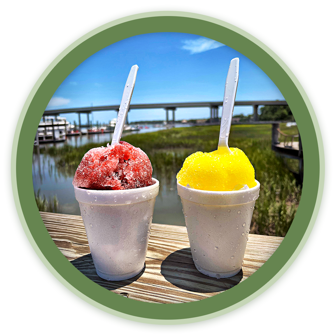 Two cups of snow cones, one red and one yellow, with plastic spoons, outdoors near an waterway with a bridge in the background.
