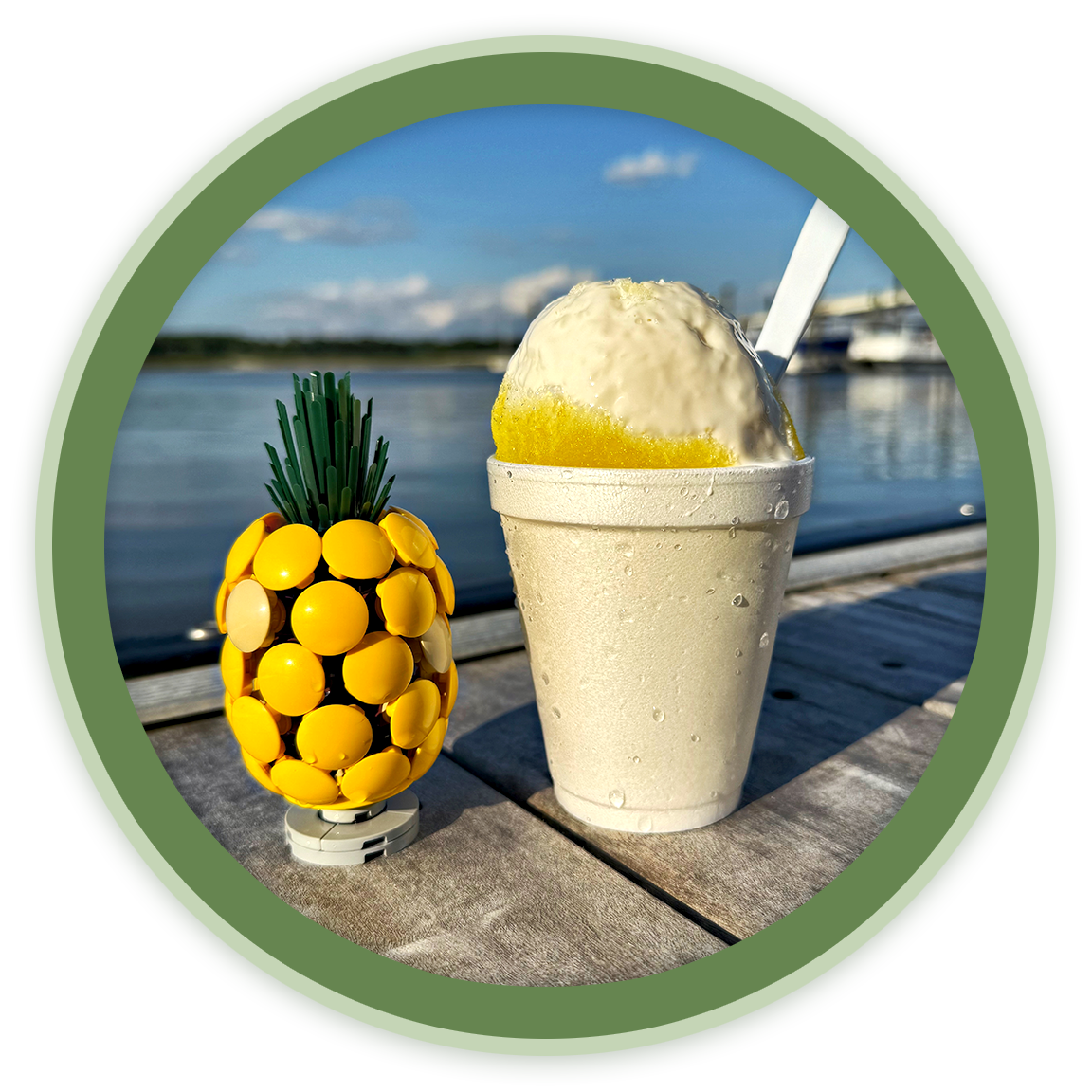 Lemon-shaped yellow object with green top next to a cup of lemonade with ice and a straw, on a wooden table by the water with boats in the background.