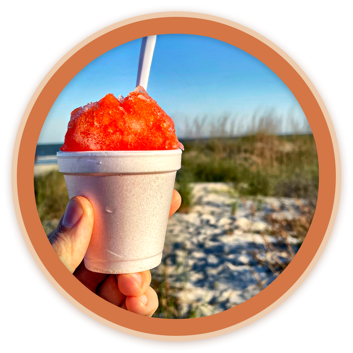 Person holding a cup of red shaved ice with a white straw on a beach with grass and sand.