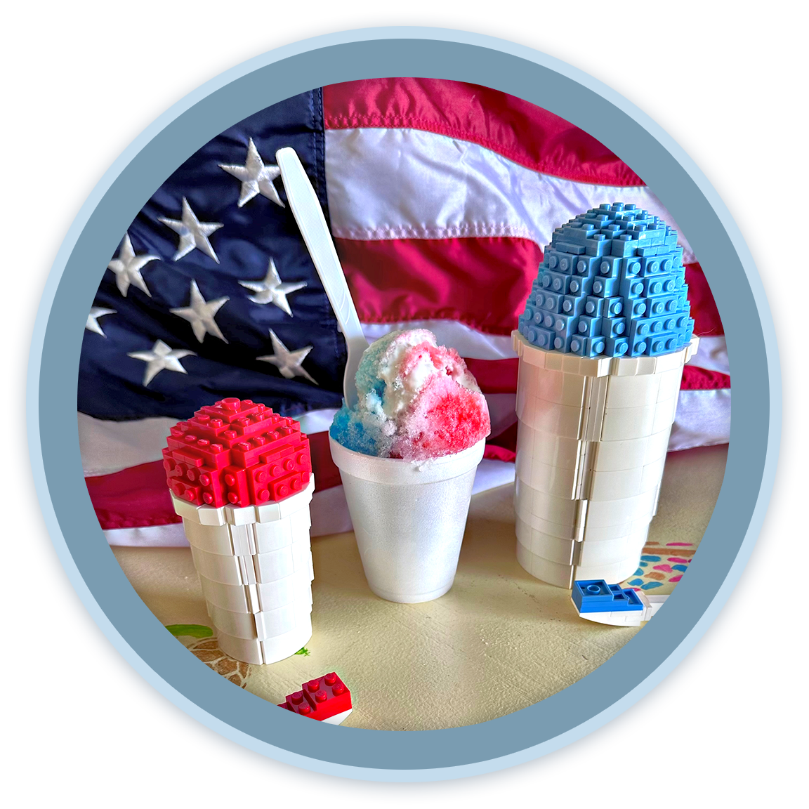 A patriotic American flag in the background, with a cup of red, white, and blue shaved ice and two ice towers made of Lego bricks, one red and one blue, on a decorated table.