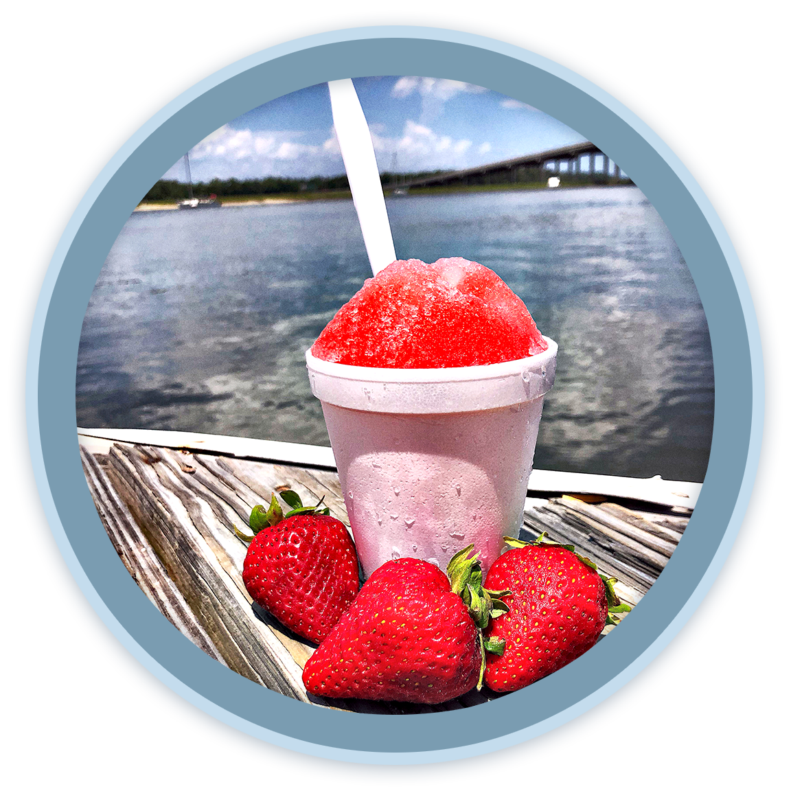 A cup of strawberry-flavored shaved ice with a red syrup on top, placed on a wooden surface next to several fresh strawberries, with a body of water and a bridge in the background.