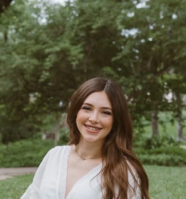 Young woman with long brown hair smiling outdoors, wearing a white top, in a green park with trees.