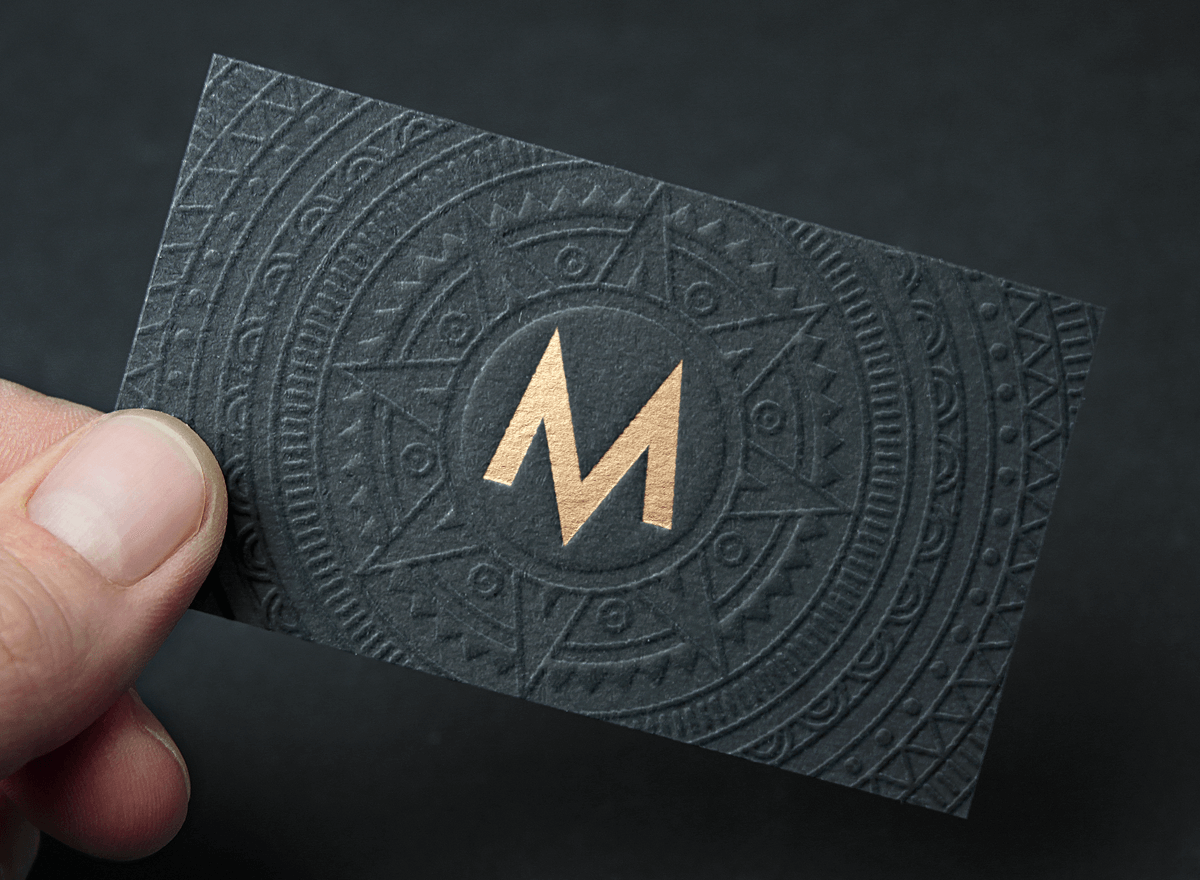 A person's hand holding a black embossed business card with gold metallic logo and intricate geometric patterns and symbols.