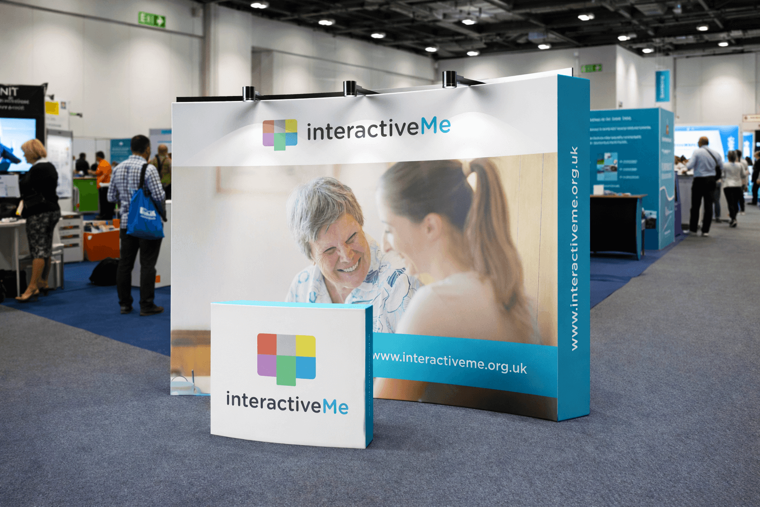 Exhibition booth for interactiveMe with a large banner showing two smiling women, one elderly and one young, engaging in conversation. The booth has a smaller sign with the interactiveMe logo, website URL www.interactiveme.org.uk, and is set up in a 