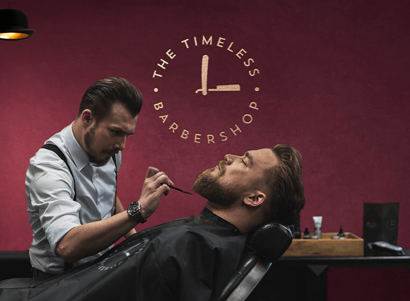Barber performing a haircut grooming service on a customer in a modern barbershop with a maroon wall and logo reading 'The Timeless Barbershop' in the background.